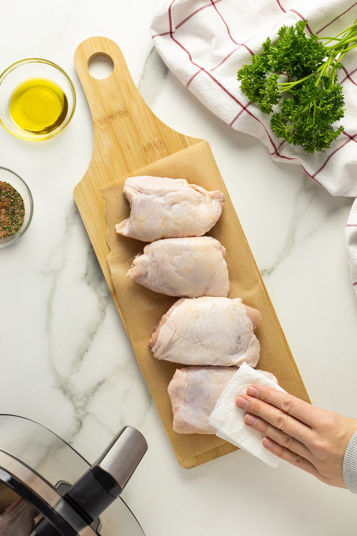 Four raw chicken thighs on a cutting board are being patted dry with a paper towel. Fresh parsley, a bowl of oil, seasoning, and a checkered kitchen towel are nearby on a marble countertop.