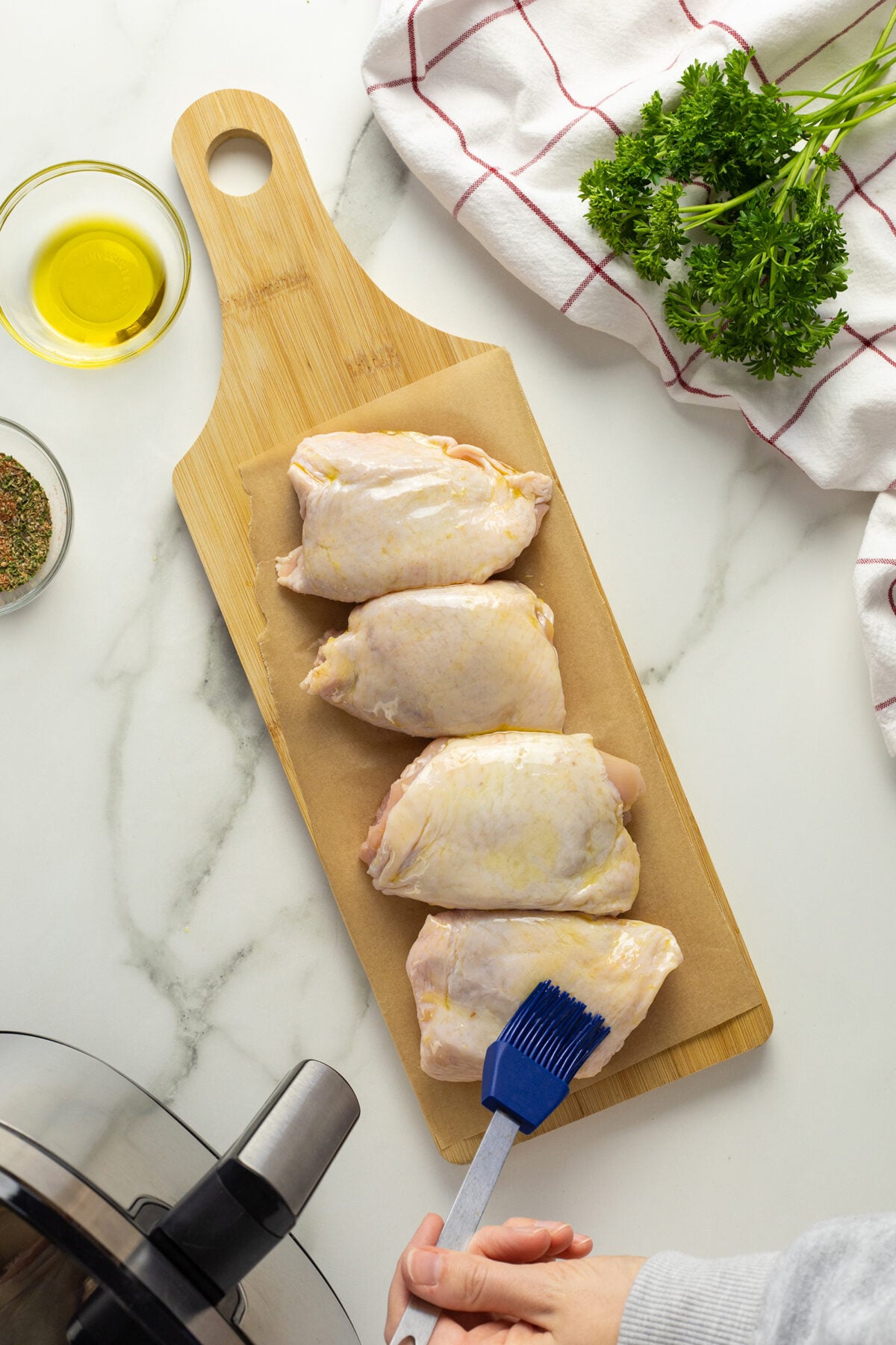 Four raw chicken thighs are lined on a wooden cutting board covered with parchment paper. A hand uses a blue brush to coat them with oil. Fresh parsley, a small bowl of oil, and a towel are nearby on the counter.