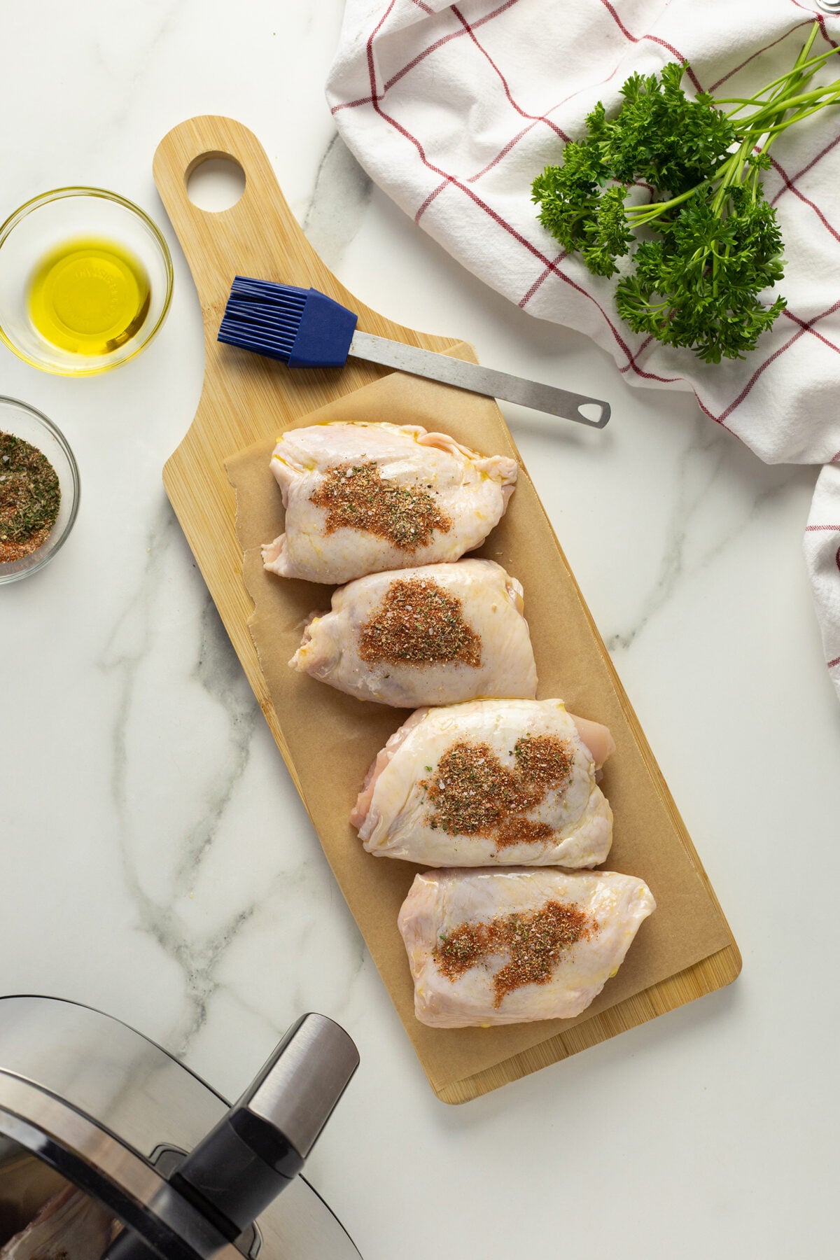 Four raw, seasoned chicken thighs are arranged on a wooden cutting board with a basting brush, next to a bowl of olive oil, fresh parsley, and a red-striped kitchen towel on a marble countertop.