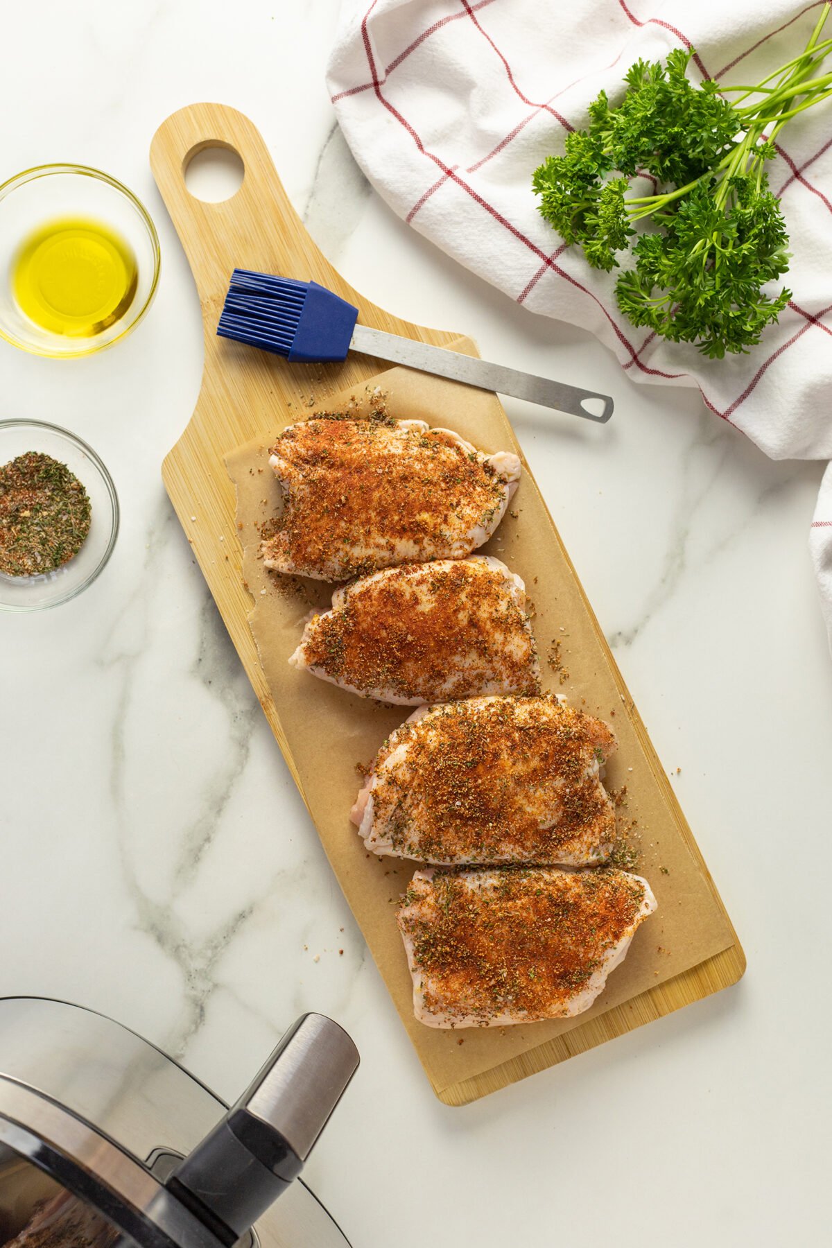 Four raw, seasoned pork chops on a wooden cutting board with a blue basting brush, fresh parsley, a bowl of oil, spice mix, and a white towel with red stripes on a marble surface. An air fryer is partially visible.