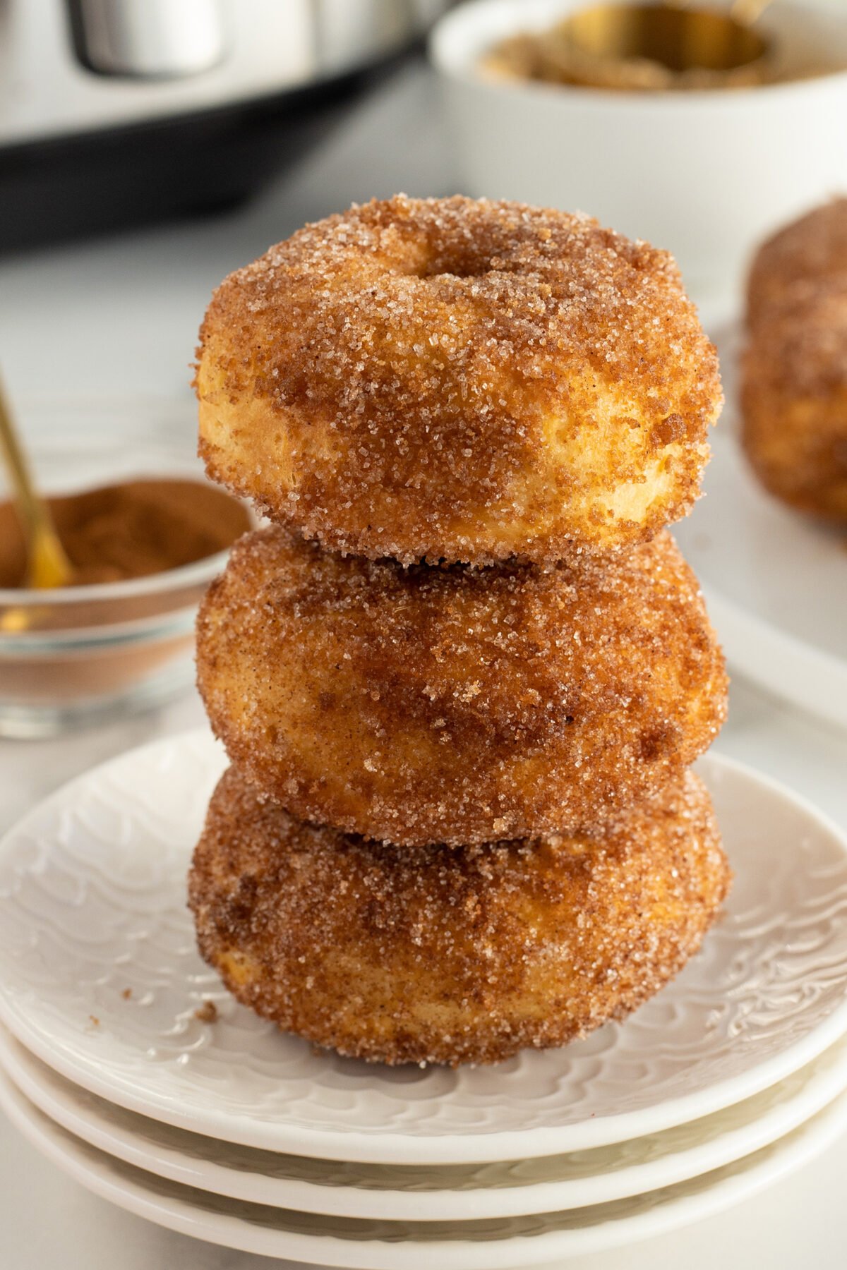 A stack of three cinnamon sugar-coated donuts sits on a white plate, with more donuts and a bowl of cinnamon in the background.