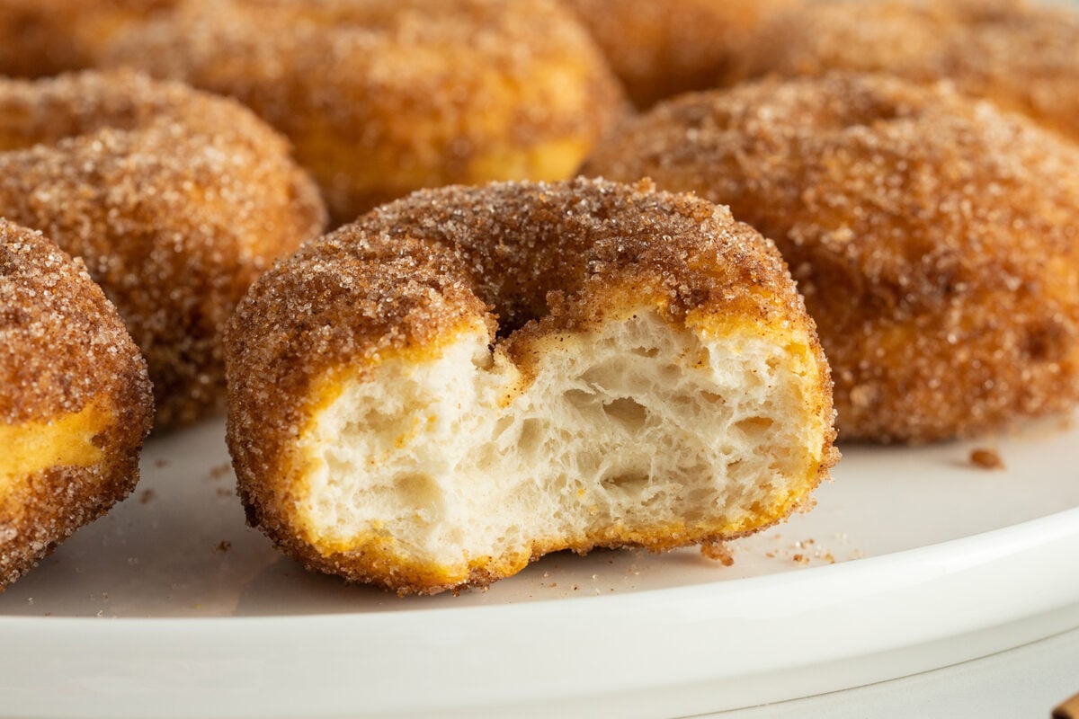 A close-up of a cinnamon sugar donut with a bite taken out, revealing its soft, airy inside. Several whole donuts are blurred in the background on a white plate.