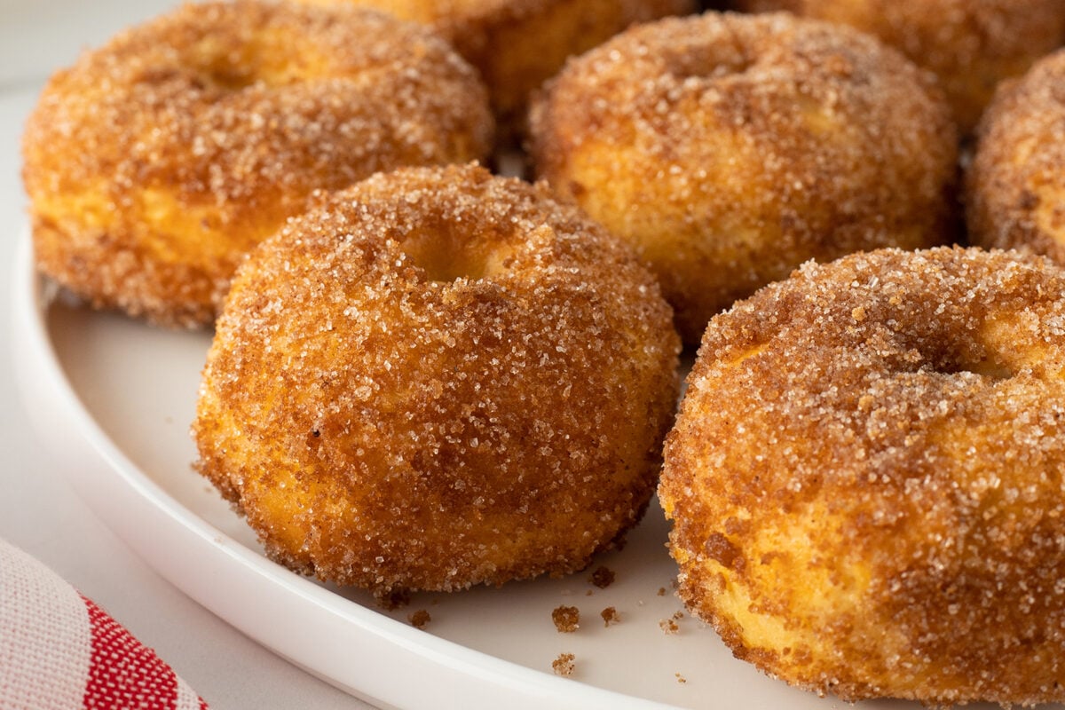 A close-up of several baked donuts covered in a generous layer of cinnamon sugar, arranged on a white plate. The donuts appear golden brown and fluffy.