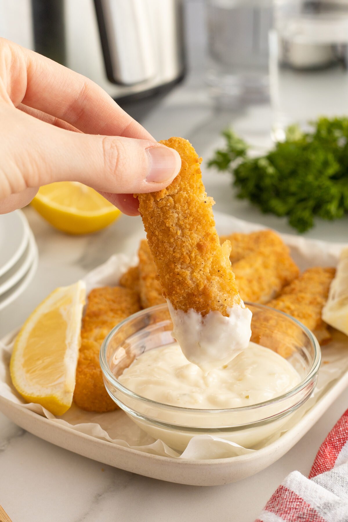 A hand dips a golden, breaded fish stick into a small glass bowl of creamy white sauce. More fish sticks and lemon wedges are on a plate in the background, with fresh parsley nearby.