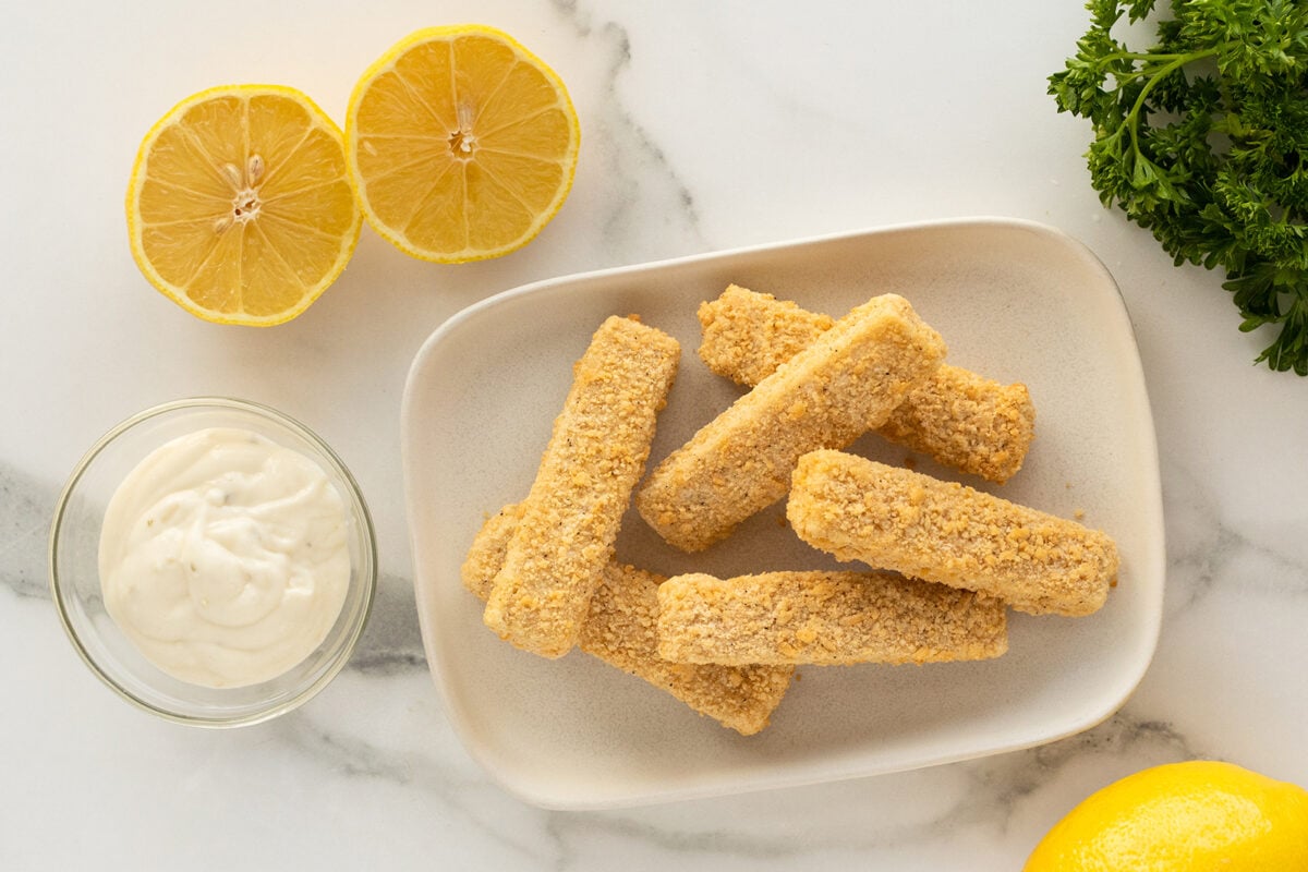 A plate of breaded fish sticks sits on a white surface, next to a small bowl of tartar sauce, two lemon halves, fresh parsley, and part of a whole lemon.