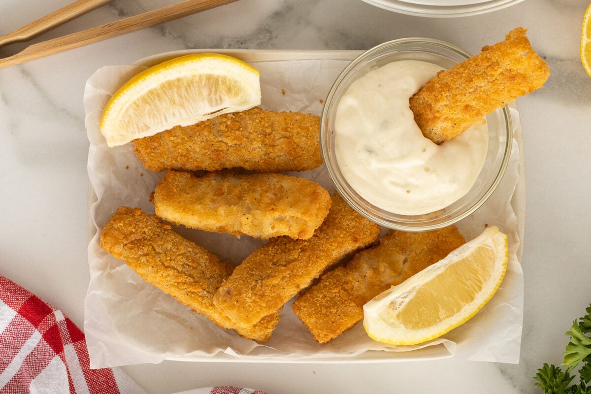 Several breaded fish sticks are arranged on parchment paper with two lemon wedges, next to a small bowl of creamy dipping sauce. One fish stick is being dipped into the sauce. A red and white cloth is partially visible.