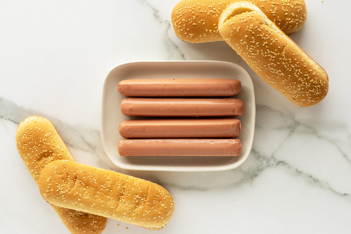 Four hot dog sausages are arranged in a rectangular dish, surrounded by four sesame seed hot dog buns on a white marble surface.