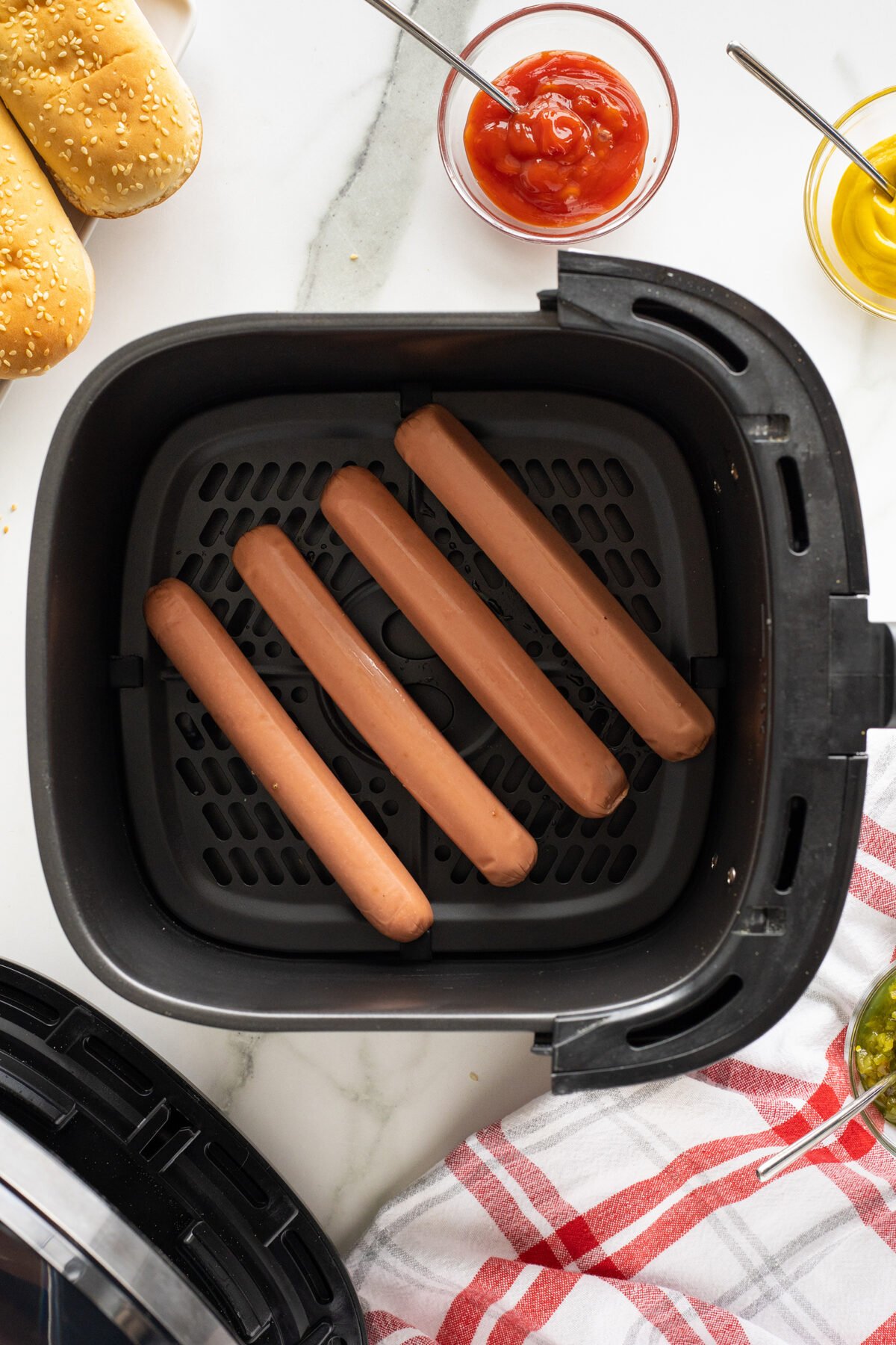 Four hot dogs arranged in the basket of an air fryer, with hot dog buns, ketchup, mustard, relish, and a red and white towel nearby on a marble surface.