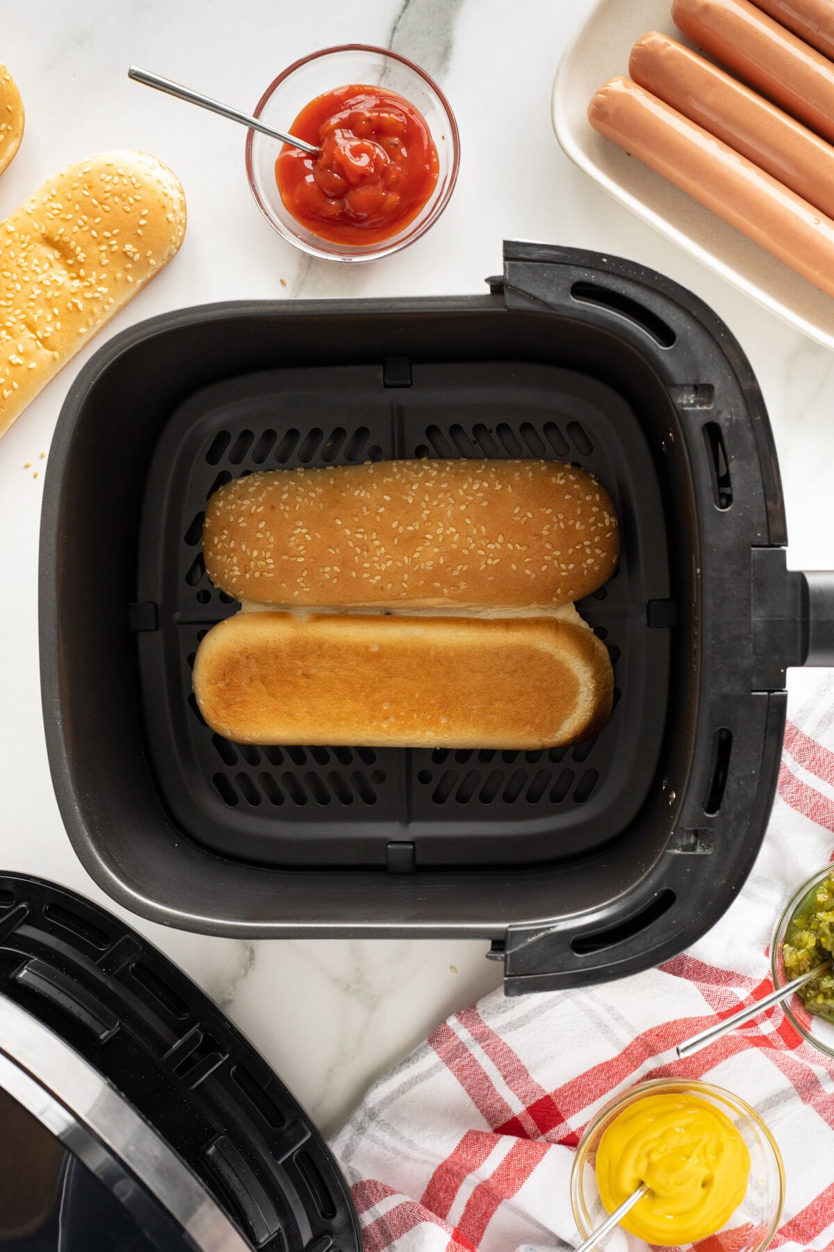 Two hot dog buns are toasting inside an air fryer basket. Nearby are bowls of ketchup and mustard, a plate with hot dogs, sliced buns, and a red and white kitchen towel on a marble countertop.