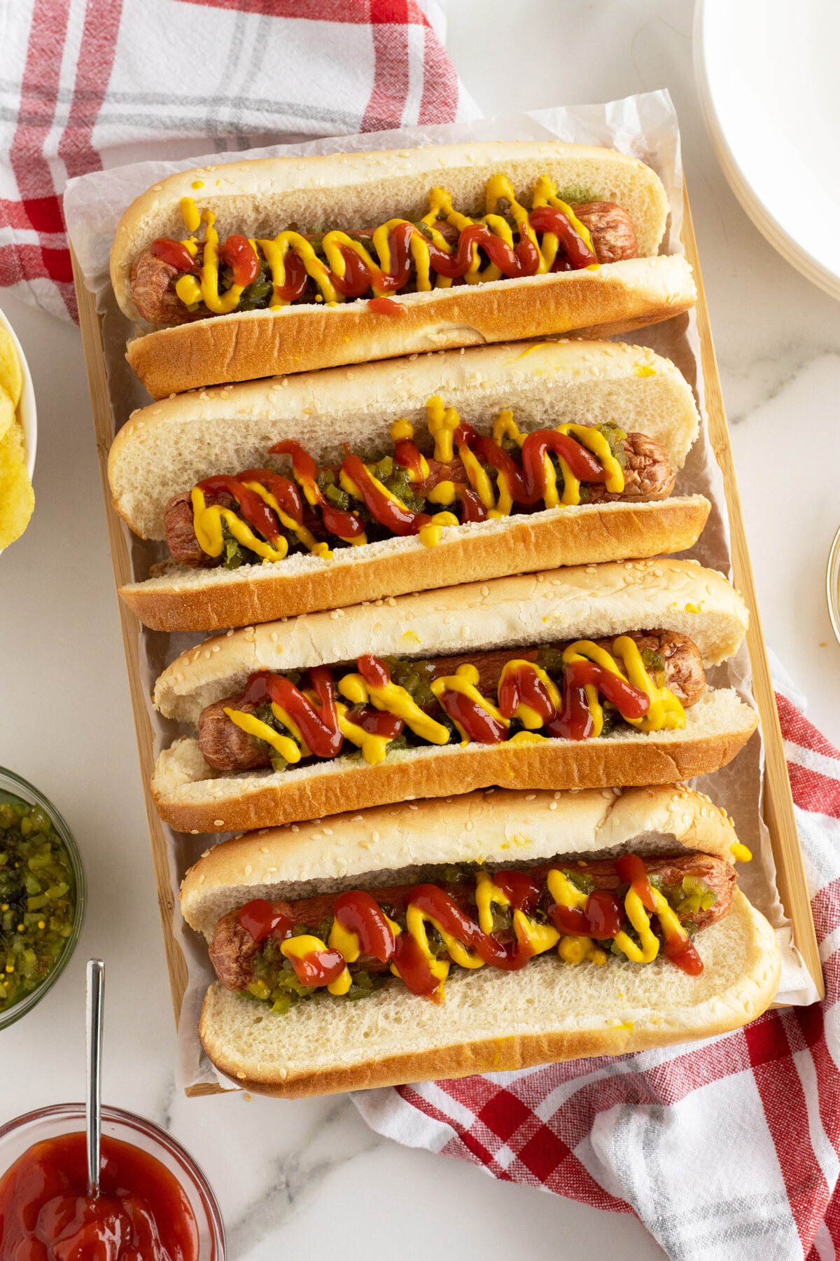 Five hot dogs in buns topped with ketchup and mustard are arranged on a tray. A jar of relish, a bowl of ketchup, plates, and a red-striped towel are nearby on a marble surface.