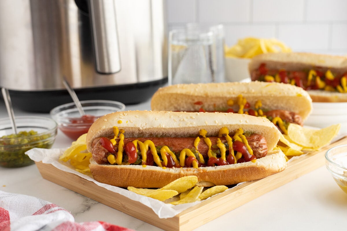 Three hot dogs topped with ketchup and mustard are served in buns on a wooden tray with potato chips. Condiment bowls with relish, ketchup, and mustard are in the background near an air fryer.