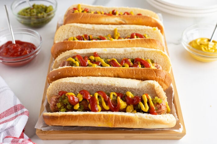 A tray of four hot dogs in buns topped with ketchup, mustard, and relish, with bowls of ketchup, mustard, and relish in the background on a white surface.