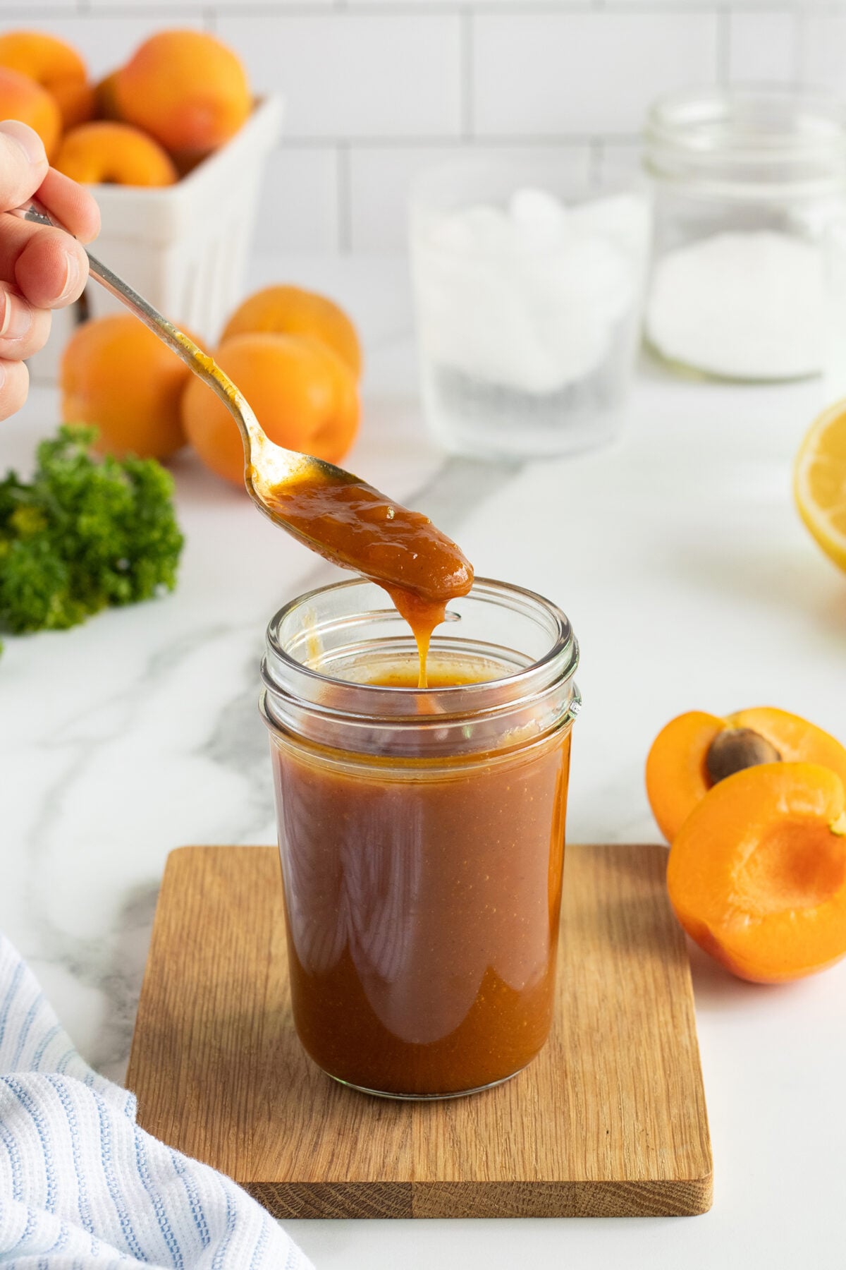 A hand holds a spoon above a jar of thick orange sauce on a wooden coaster. Fresh apricots, a sliced lemon, and green herbs are in the background on a white countertop.