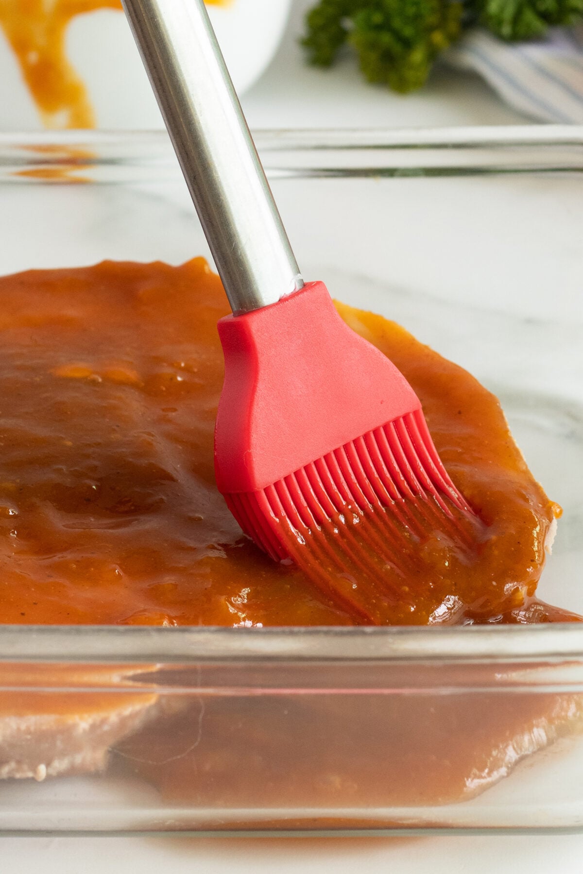 A red silicone basting brush is spreading a thick, orange-brown sauce over a piece of meat in a glass dish.