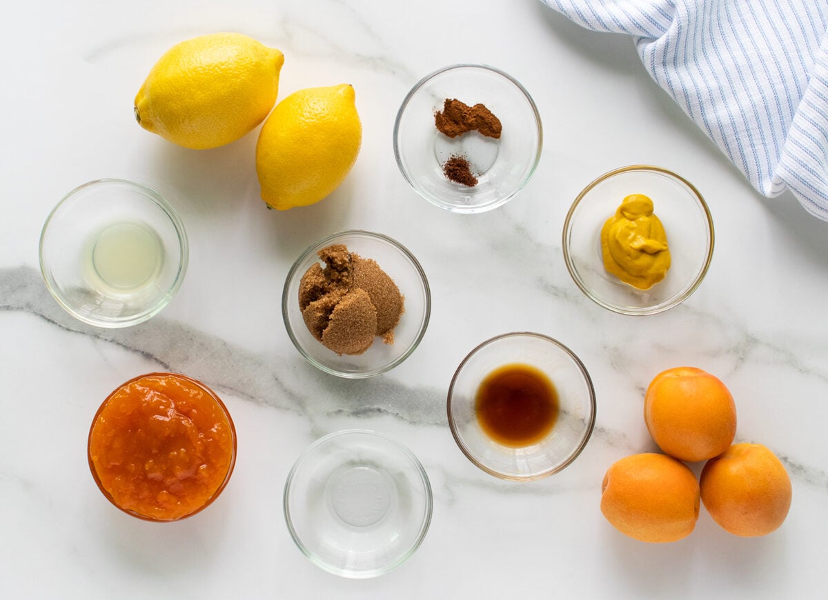 Glass bowls with brown sugar, mustard, apricot jam, lemon juice, spices, and vanilla extract are arranged on a white marble surface, with whole lemons and apricots and a striped cloth visible in the corners.