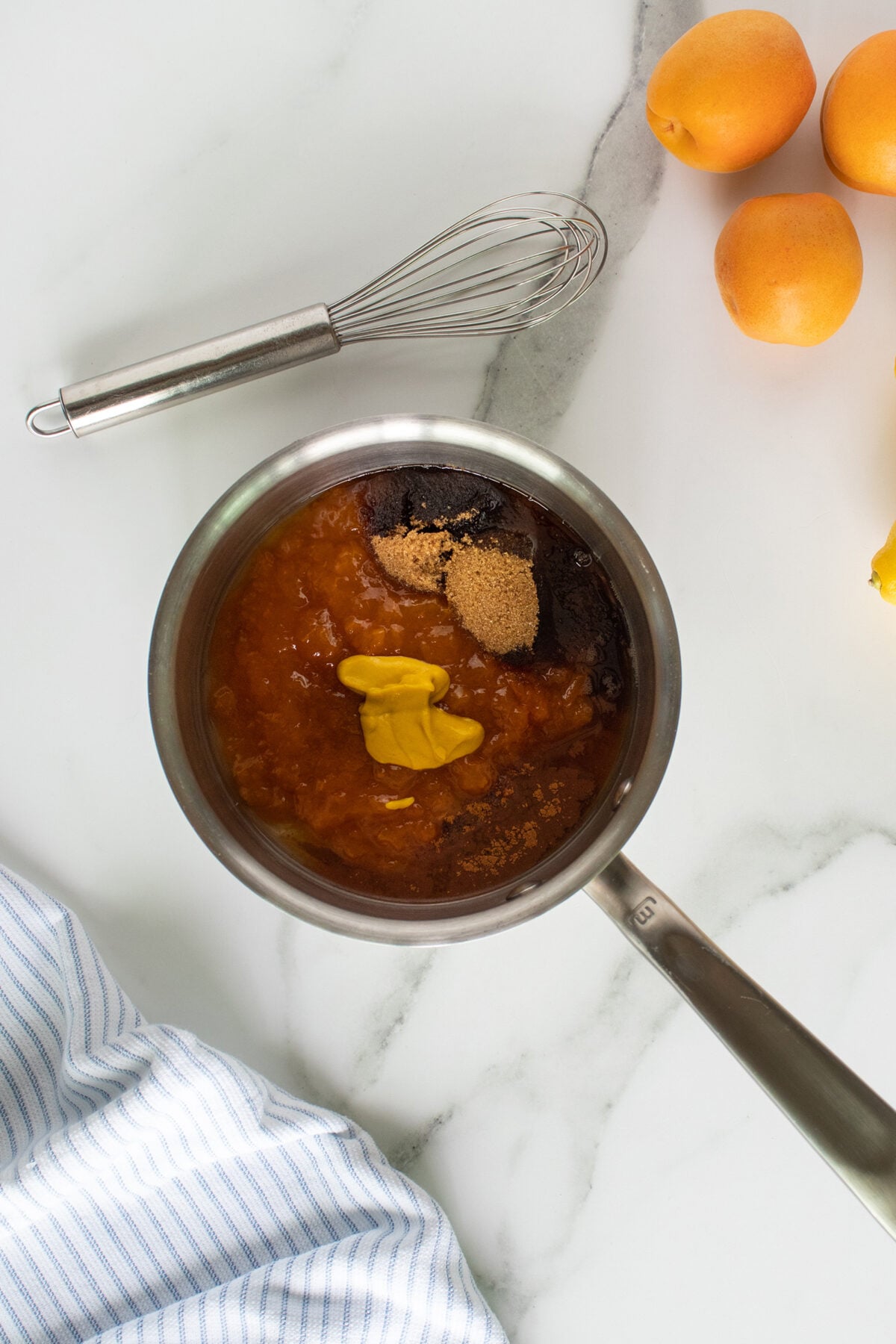 A saucepan with apricot sauce, brown sugar, spices, and mustard on a marble counter, next to a whisk, fresh apricots, and a striped kitchen towel.