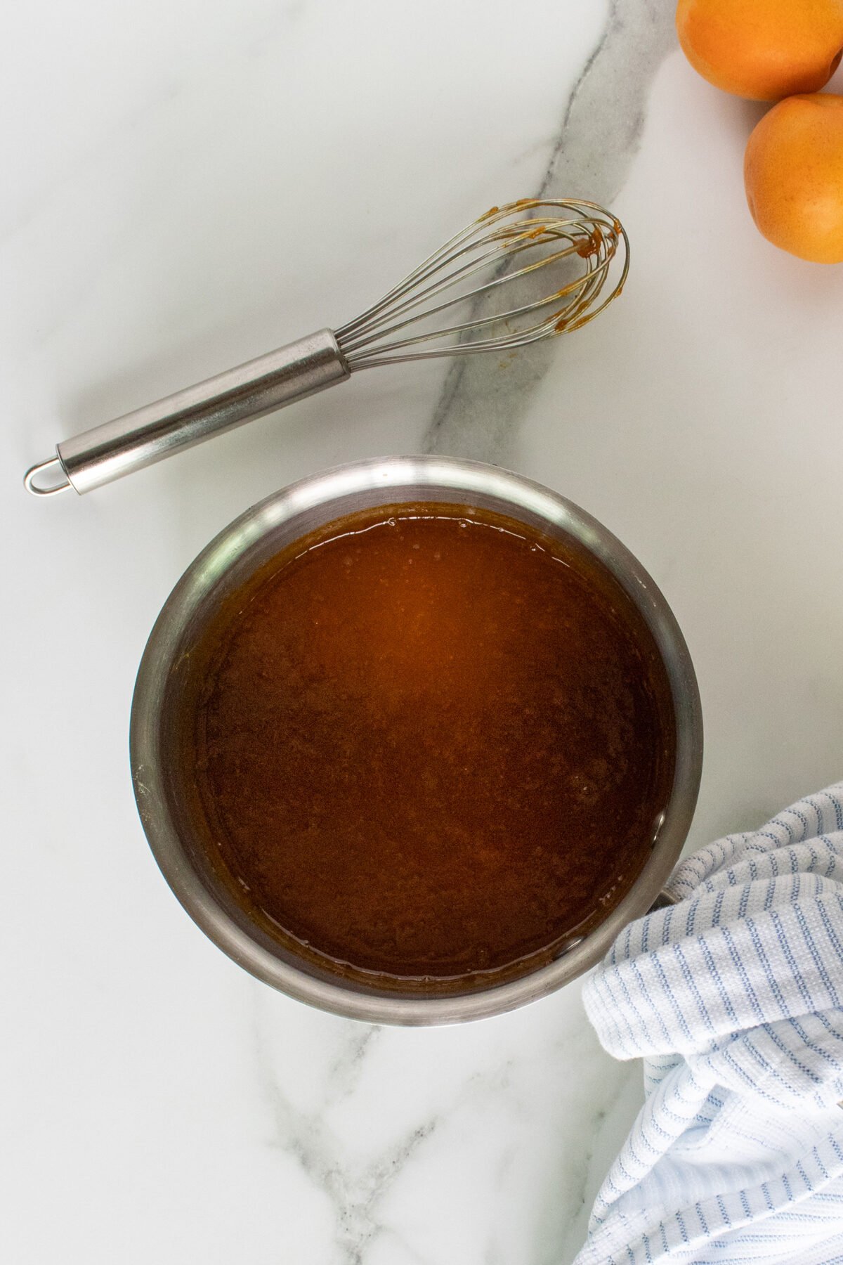 A metal bowl filled with a golden-brown liquid sits on a marble surface next to a whisk with some residue, a blue-and-white striped towel, and a few orange apricots in the corner.