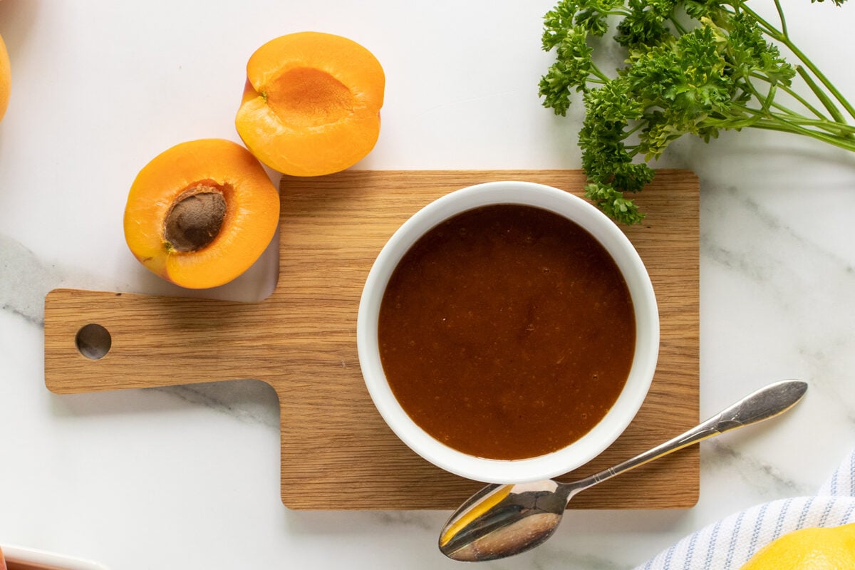 A small bowl of brown sauce with a spoon sits on a wooden board, next to a halved apricot, a whole apricot, and a bunch of fresh parsley on a white surface.
