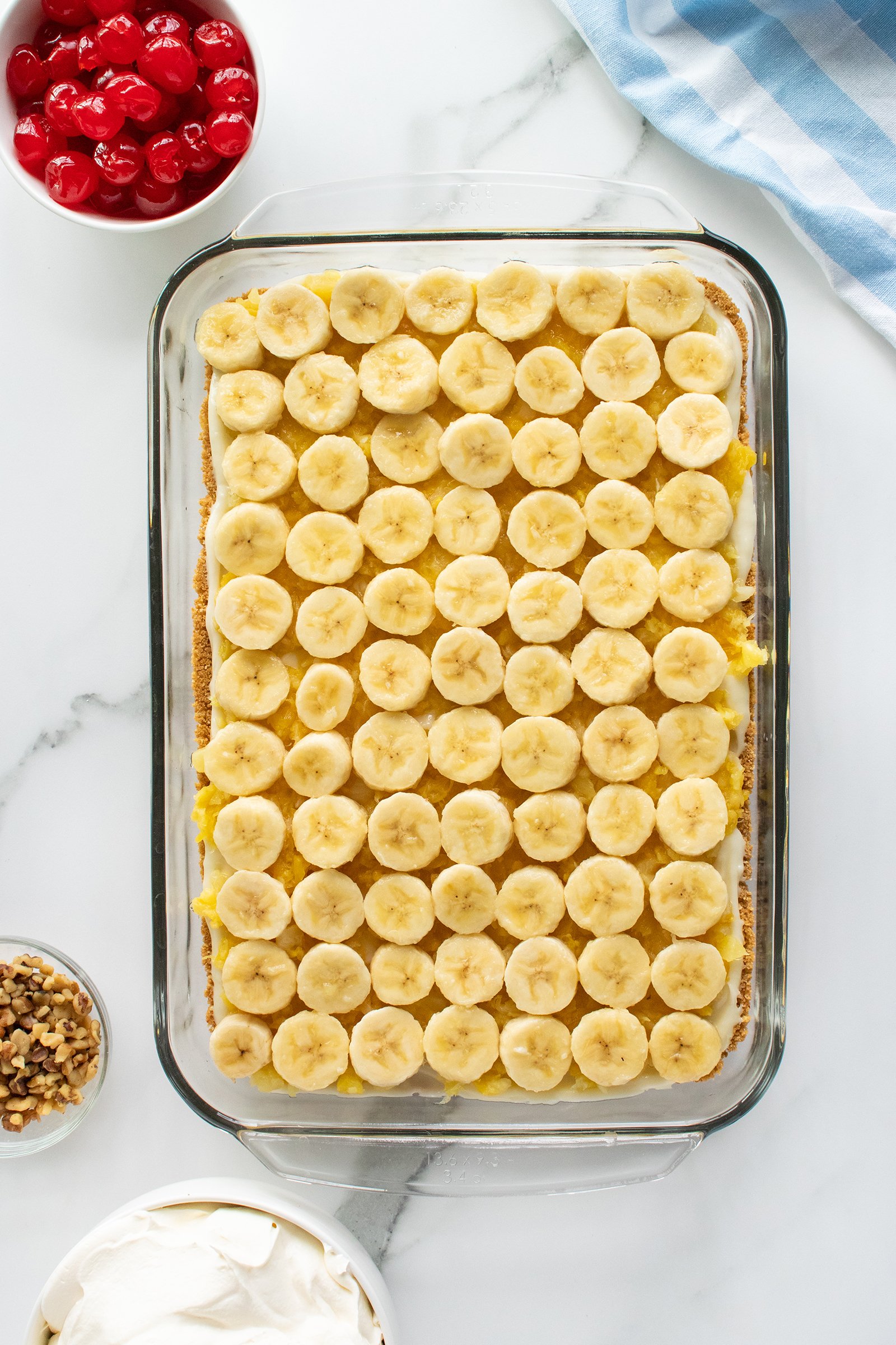 A glass baking dish filled with a dessert topped with neatly arranged banana slices. Surrounding the dish are bowls of whipped cream, chopped nuts, and maraschino cherries on a white marble surface.