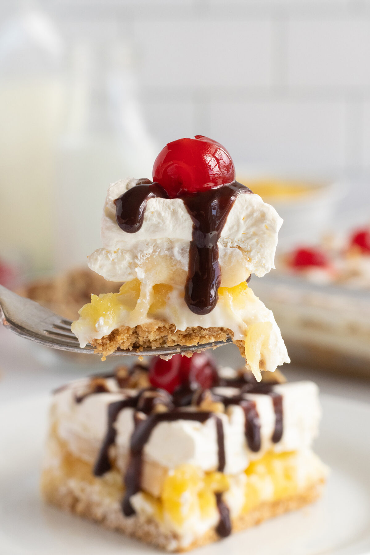 A fork holds a bite of dessert topped with whipped cream, a maraschino cherry, and chocolate drizzle. Below, a larger piece of the same layered dessert sits on a plate.