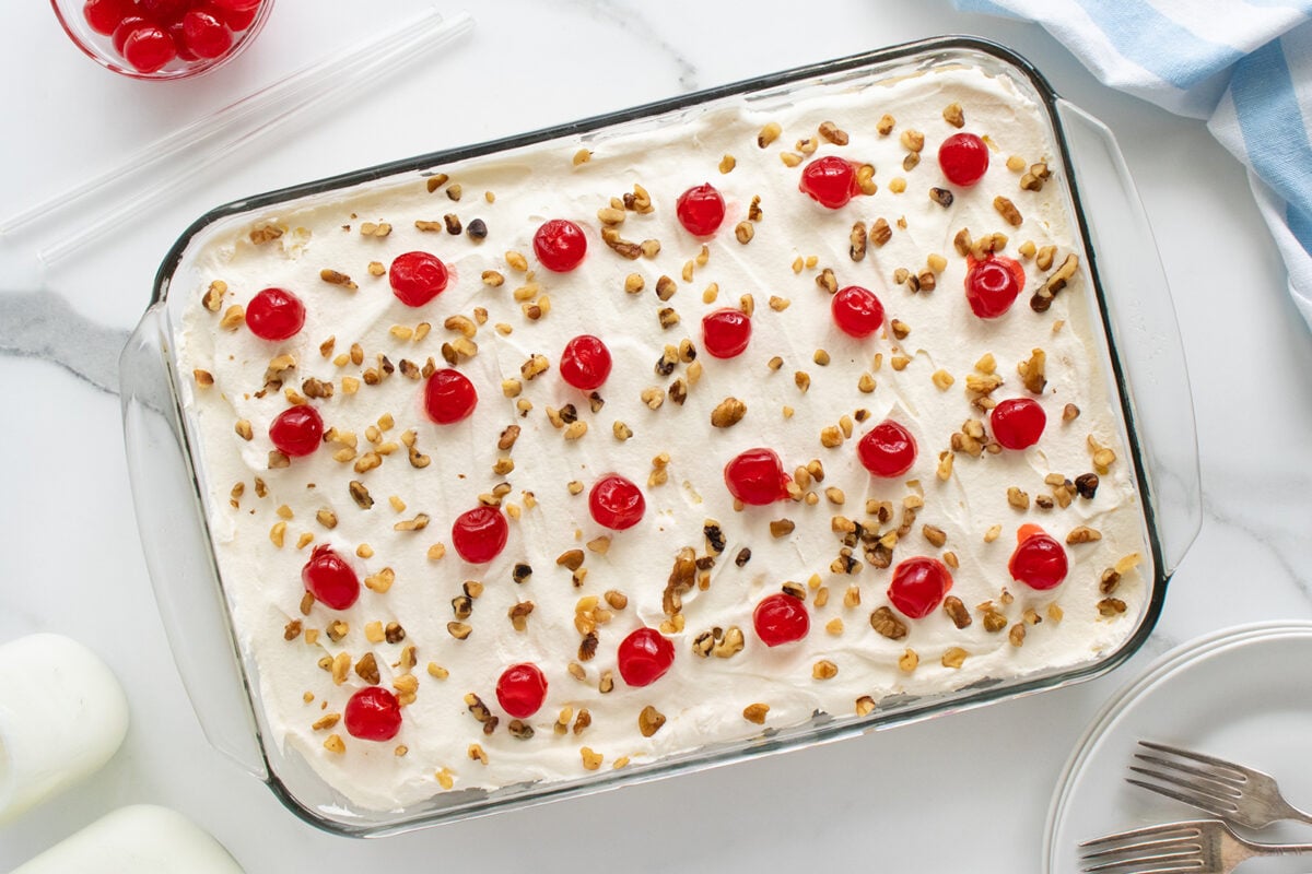 A rectangular glass dish filled with whipped cream-topped dessert, garnished with maraschino cherries and chopped nuts, sits on a white surface near plates, forks, and a bowl of extra cherries.