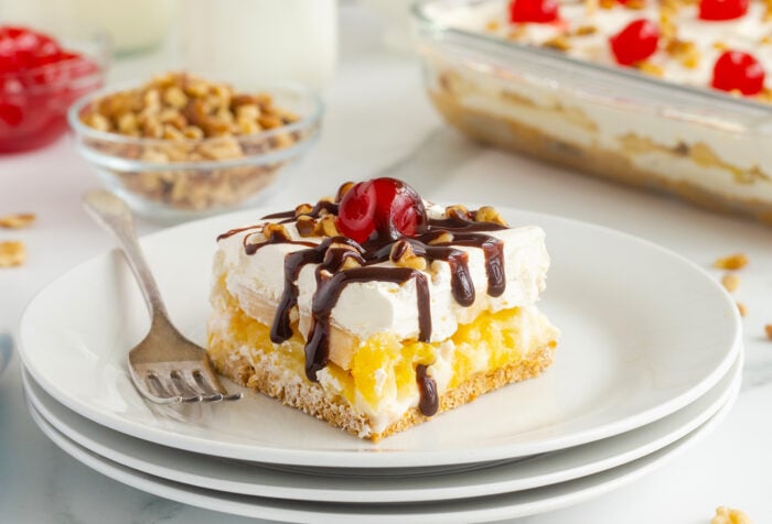 A dessert square with whipped cream, pineapple, a graham cracker crust, chocolate drizzle, chopped nuts, and a cherry on top, served on a white plate with a fork. A bowl of nuts and a dessert dish appear in the background.