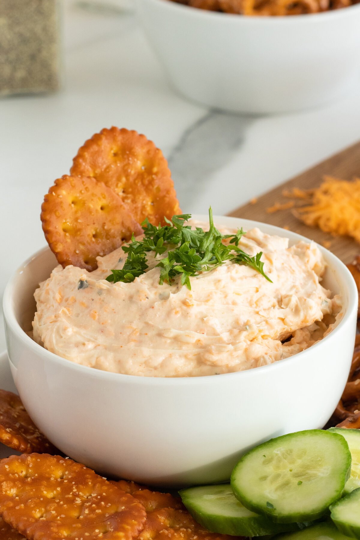 A white bowl filled with creamy dip, garnished with chopped herbs and two round crackers. Sliced cucumber and more crackers are arranged around the bowl on a wooden board.