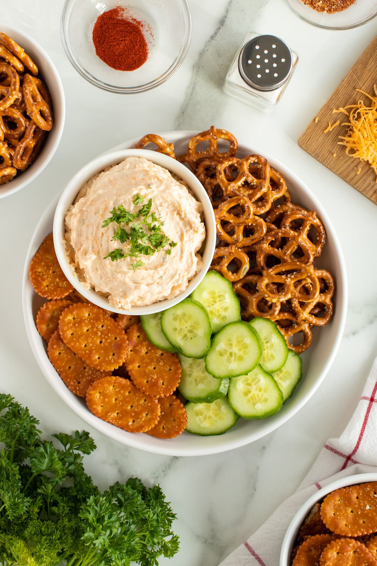 A bowl with creamy dip topped with herbs, surrounded by round crackers, cucumber slices, and pretzels. Nearby are shredded cheese, paprika, a salt shaker, and fresh parsley on a marble surface.