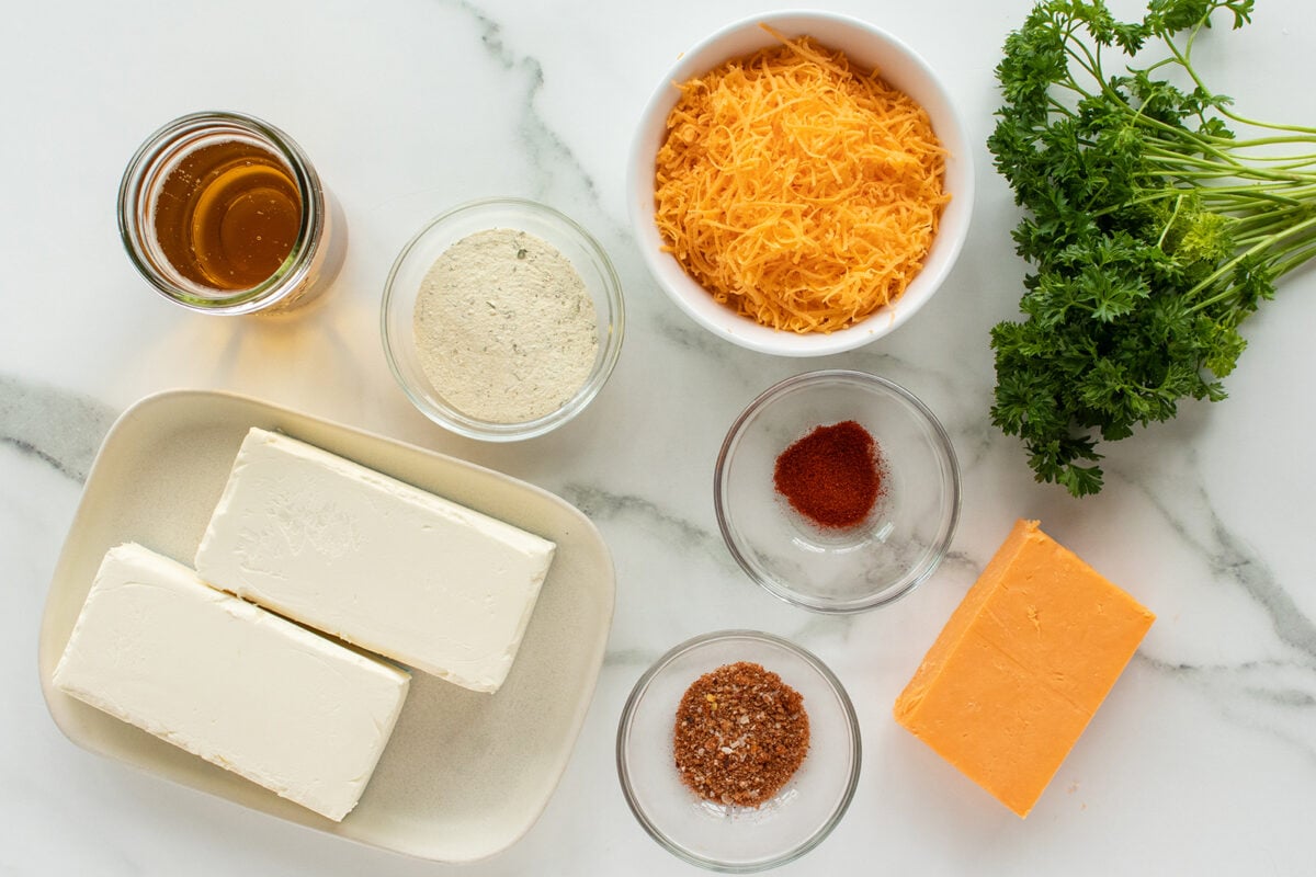 Ingredients arranged on a marble surface: two blocks of cream cheese, a jar of honey, a bowl of shredded cheddar, a bunch of fresh parsley, a block of cheddar, flour, paprika, salt, and a seasoning mix in small bowls.