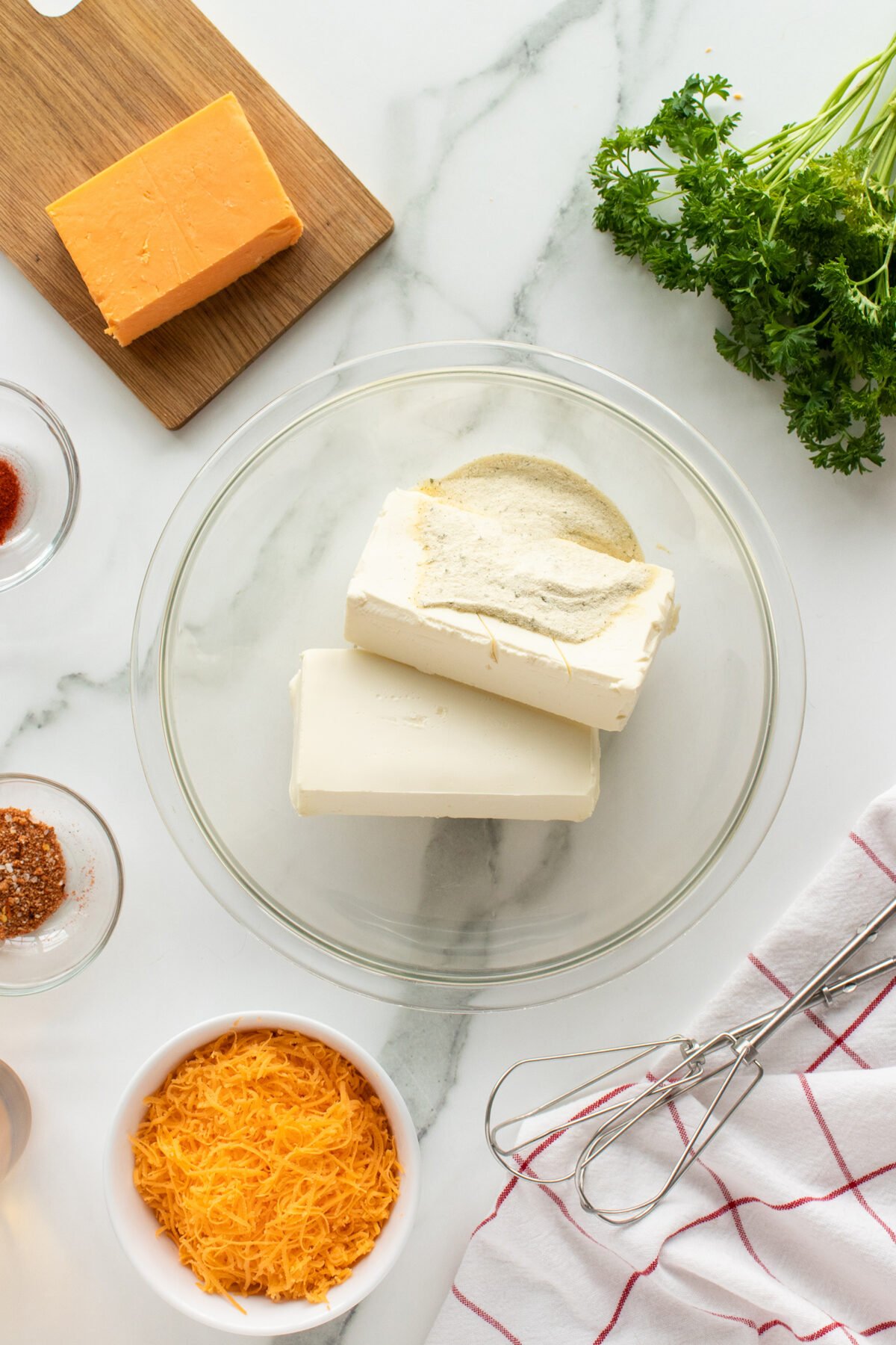 A glass bowl with cream cheese and seasonings on a marble surface, surrounded by a block and bowl of cheddar cheese, fresh parsley, spices, a whisk, and a red-striped kitchen towel.
