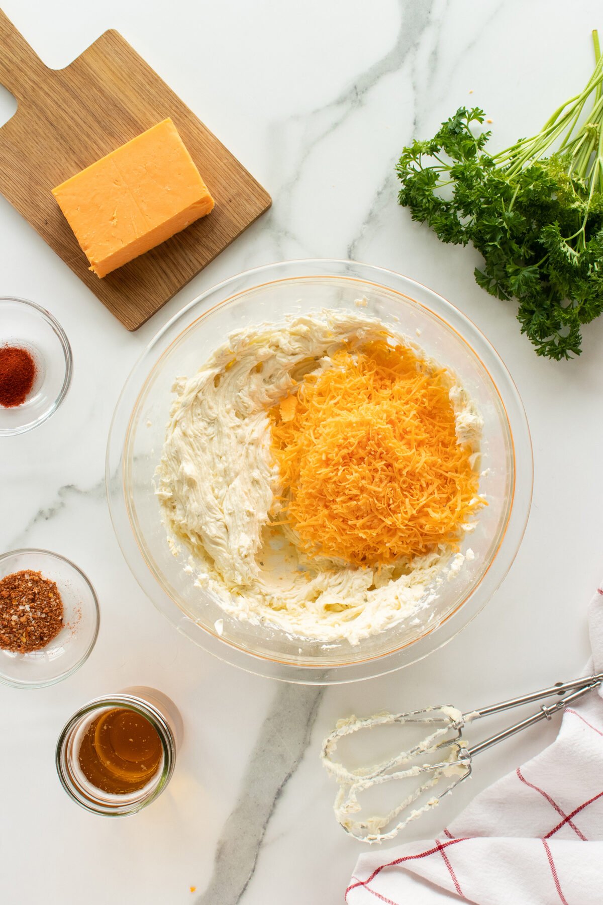 A glass bowl with cream cheese and shredded cheddar cheese being mixed, surrounded by a block of cheddar, fresh parsley, spices, honey, and a hand mixer on a white marble countertop.