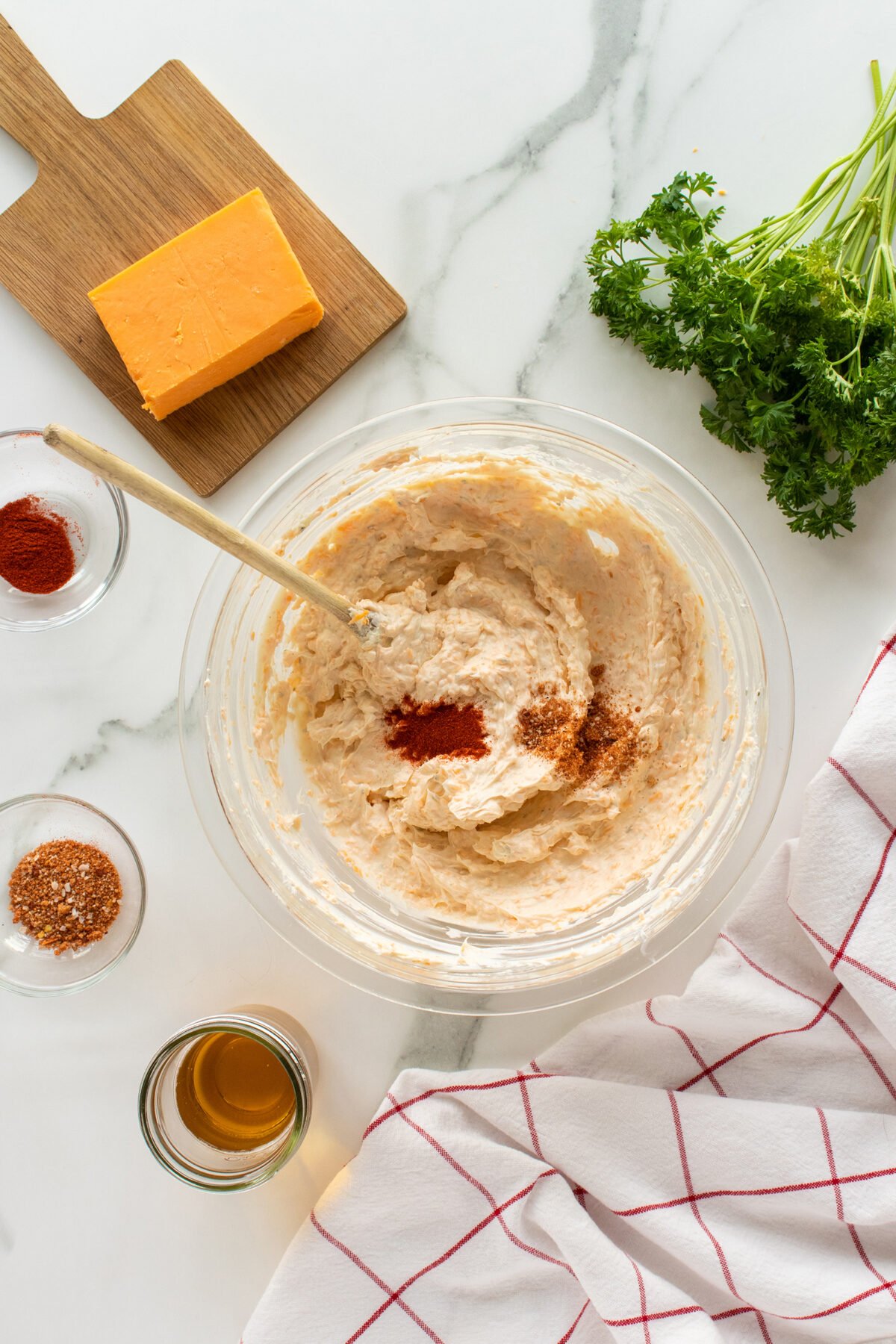 A glass bowl with a creamy mixture and spices, surrounded by a wooden board with cheddar cheese, fresh parsley, small bowls of spices and liquid, and a white towel with red stripes on a marble surface.