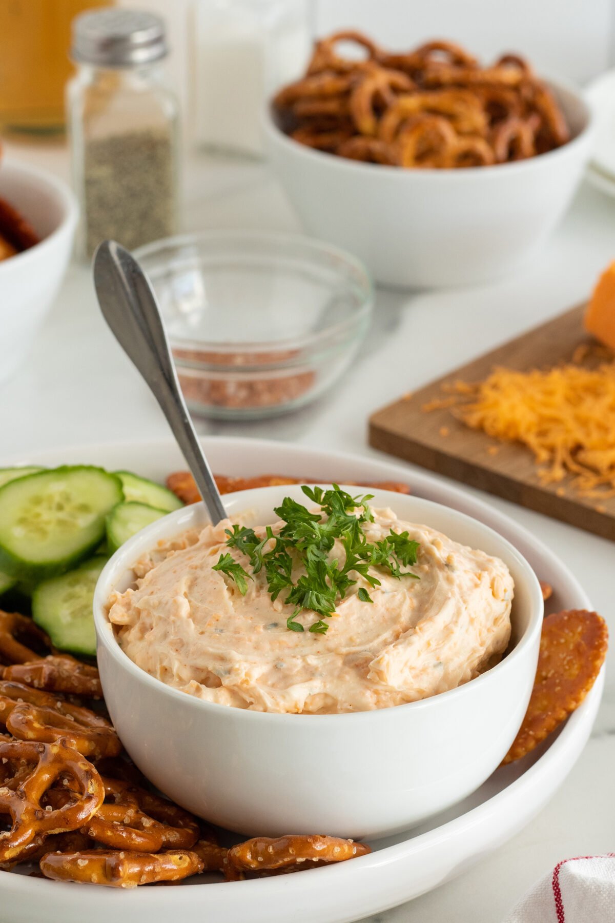 A white bowl of creamy cheese dip garnished with parsley, surrounded by pretzels and cucumber slices. Shredded cheese, a bowl of pretzels, and seasoning are visible in the background.