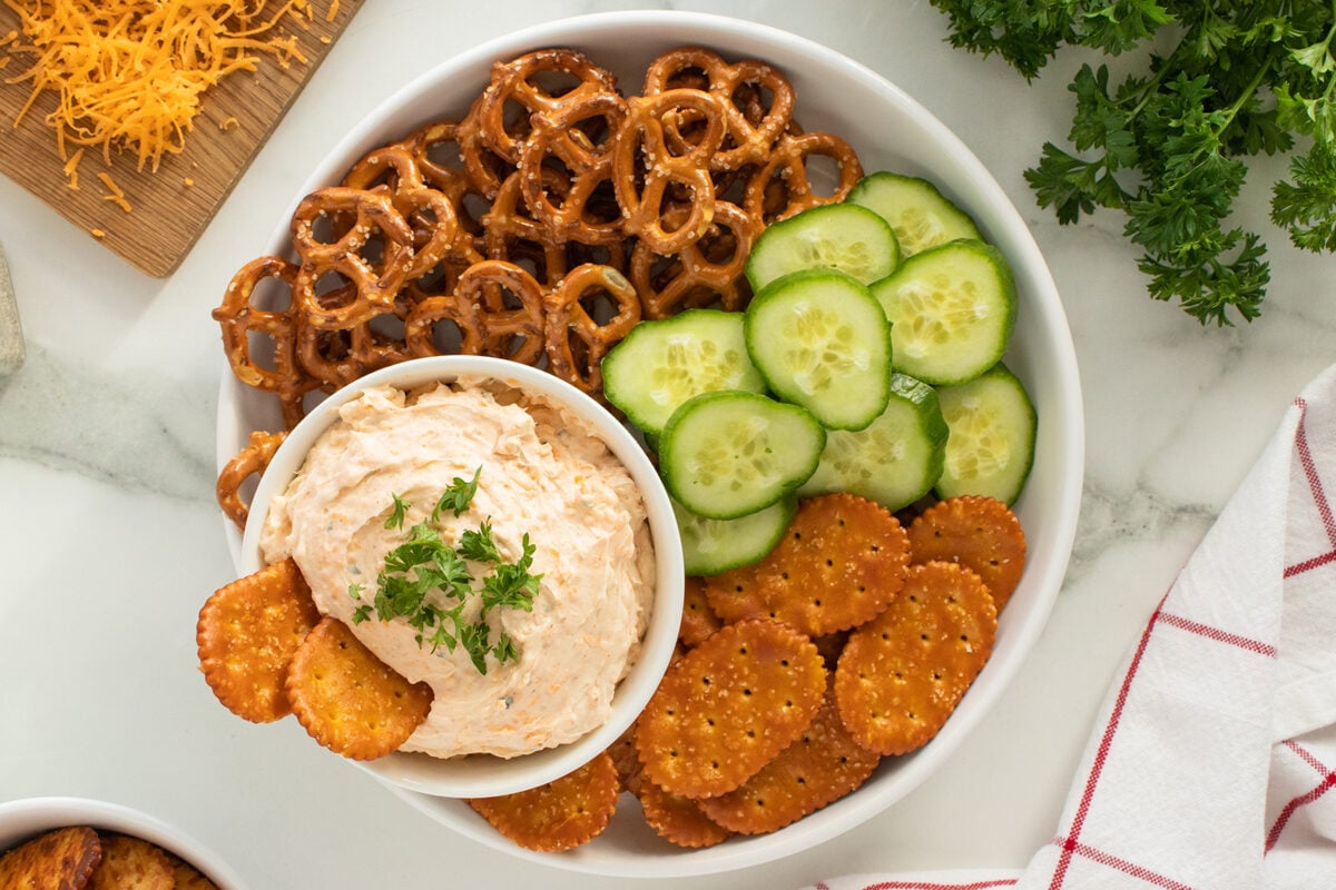 A white plate with pretzels, cucumber slices, and round crackers arranged around a bowl of creamy dip topped with herbs. A cutting board with shredded cheese and parsley are nearby.