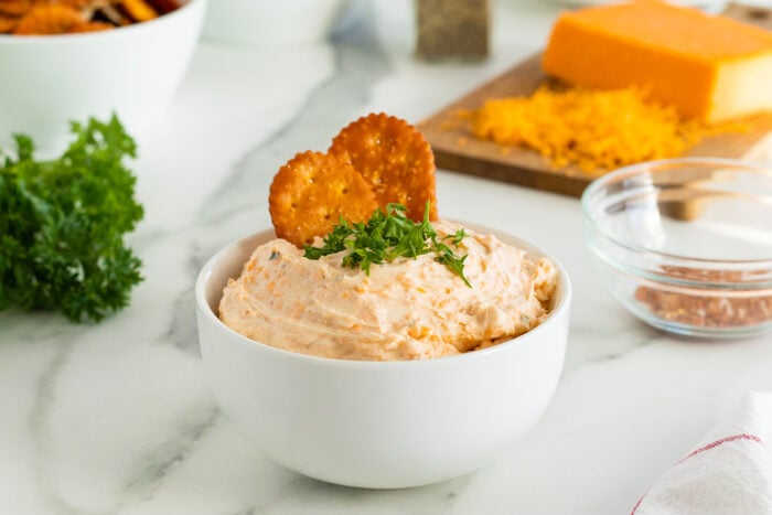 A white bowl filled with creamy cheese dip, garnished with chopped herbs and two round crackers, sits on a marble counter. In the background, there is a block of cheddar cheese and a glass bowl with seasonings.