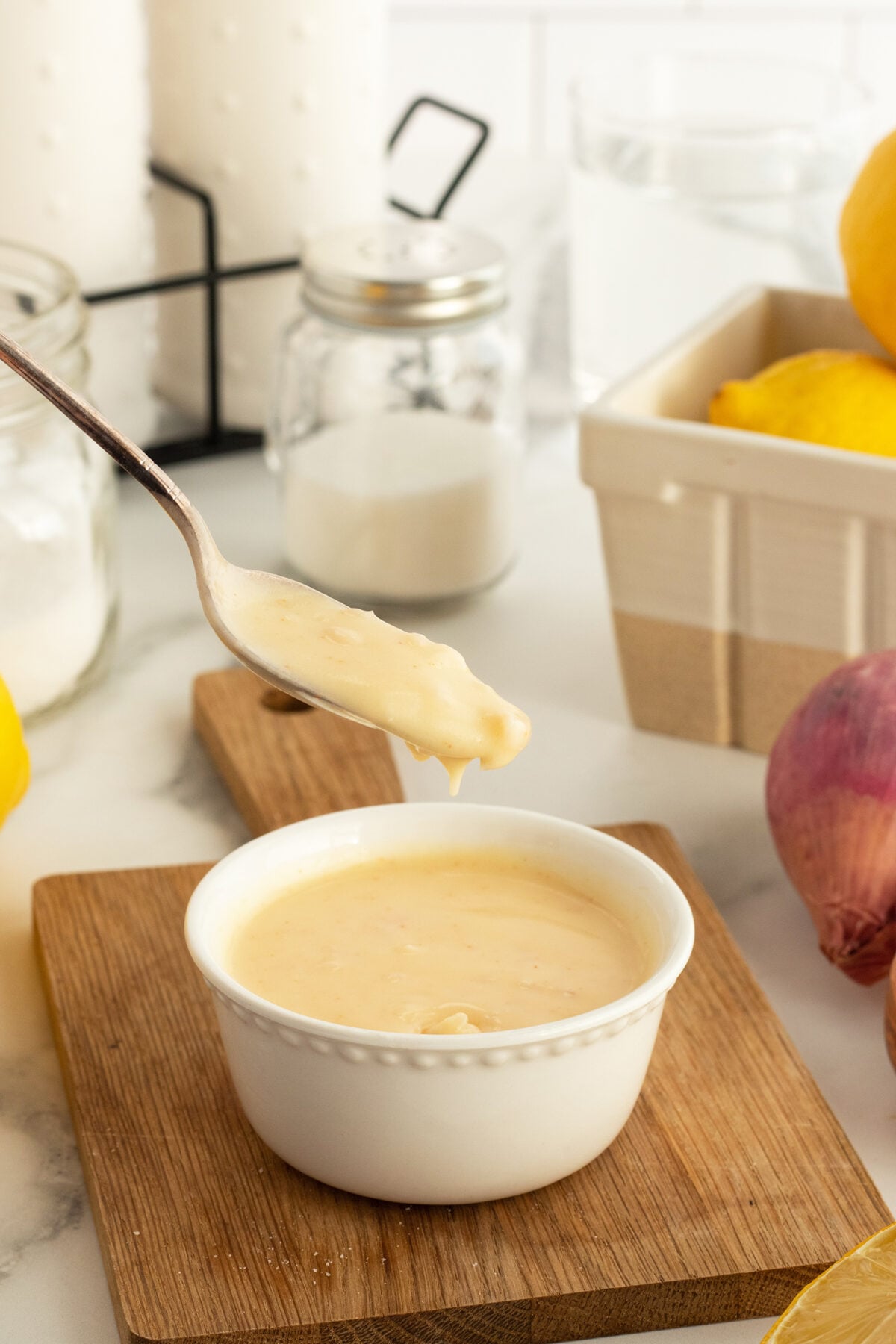 A spoon holds up a thick, creamy yellow sauce above a small white bowl on a wooden board. Lemons, a red onion, and kitchen jars are visible in the background on a white countertop.