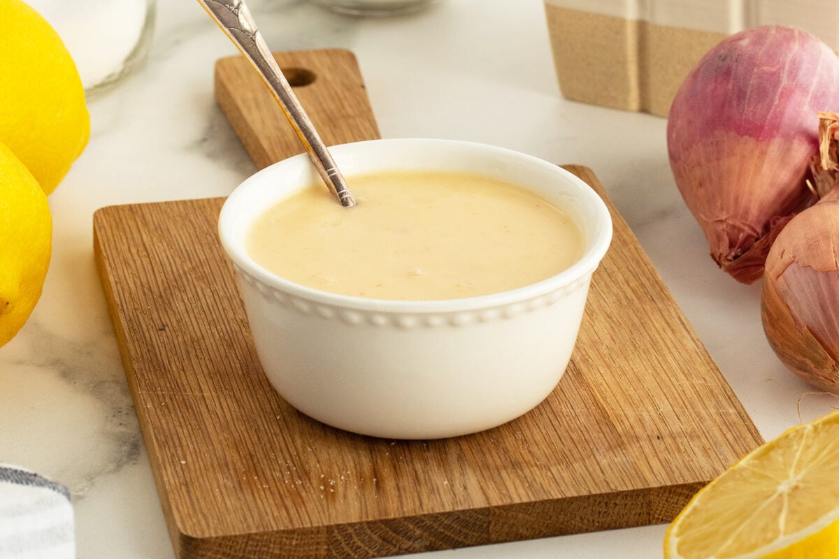 A small white bowl filled with creamy dressing sits on a wooden board, with a spoon inside. Fresh lemons and shallots are nearby on a marble countertop.