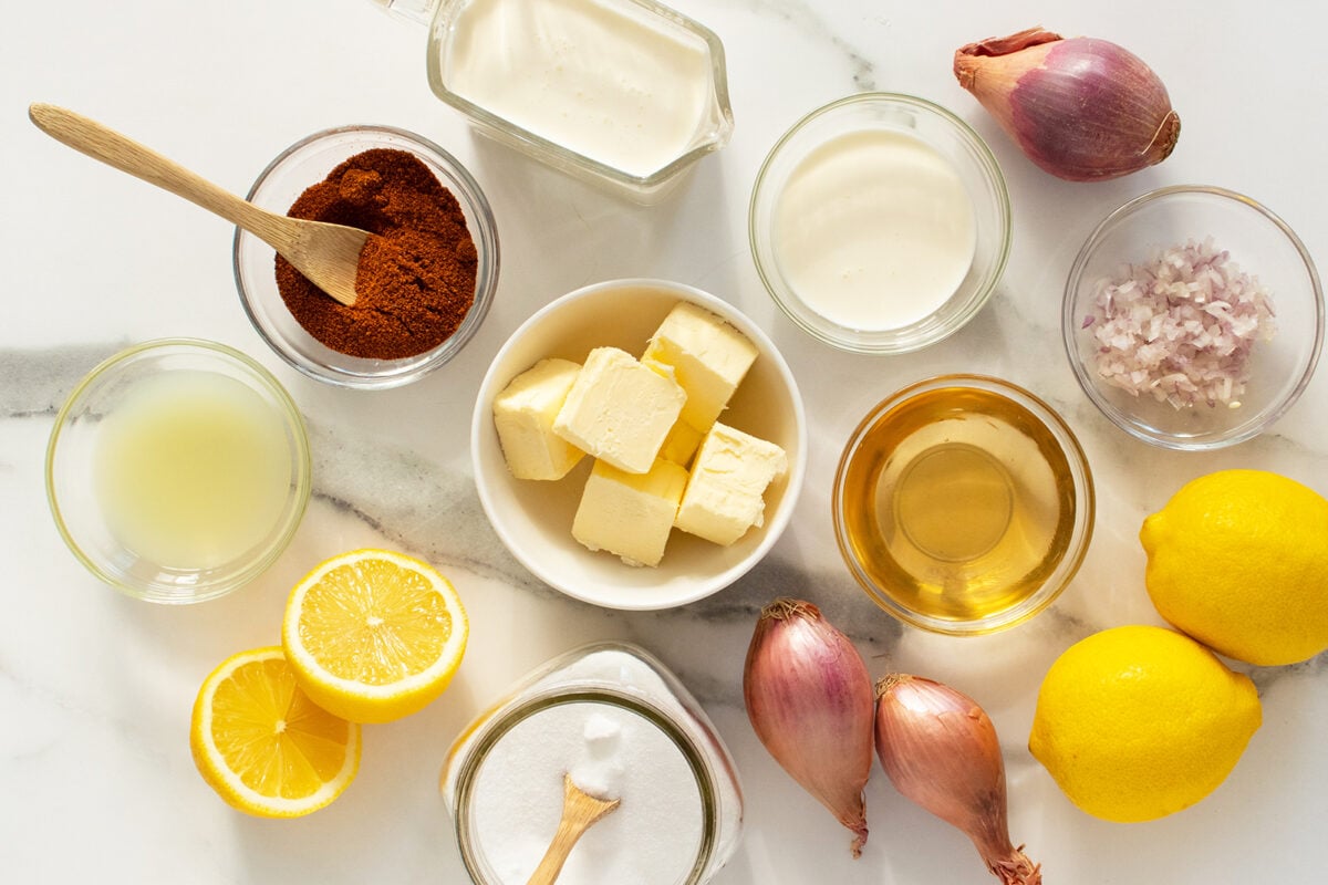 An overhead view of ingredients on a marble surface, including halved lemons, whole shallots, cubed butter, cream, oil, lemon juice, salt, paprika, and finely chopped shallot in small glass bowls.