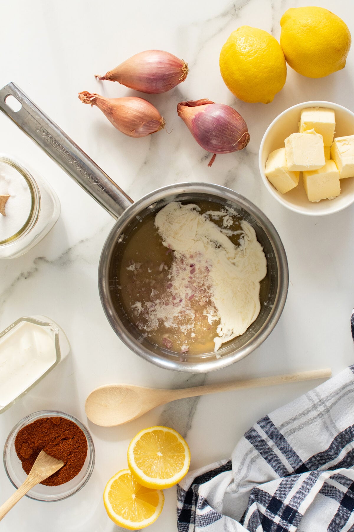 A saucepan with cream and shallots sits on a marble counter surrounded by lemons, shallots, butter cubes, paprika, a jar of cream, sugar, a wooden spoon, and a striped cloth.