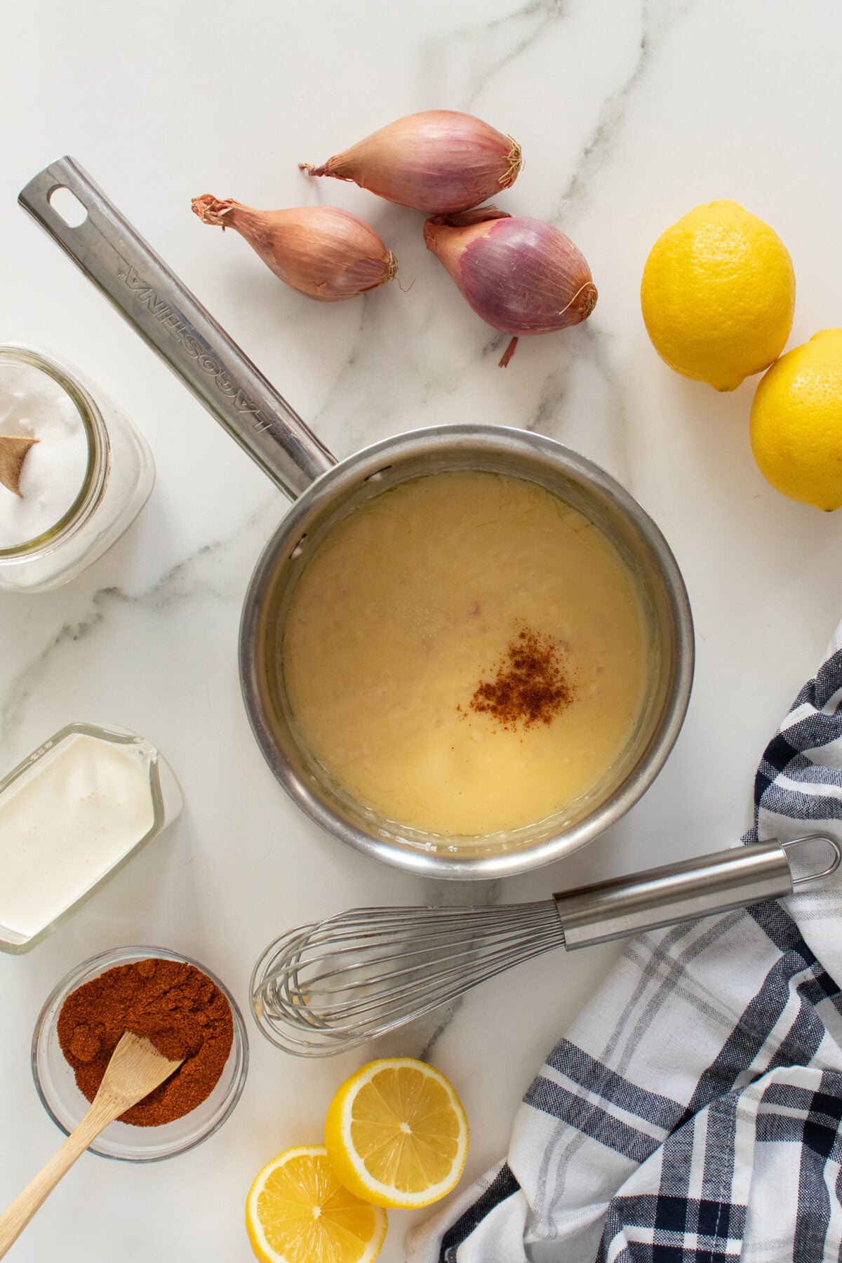 A saucepan with a creamy yellow sauce and a sprinkle of spice sits on a marble surface, surrounded by shallots, lemons, a jar of cream, ground spice, a whisk, and a striped cloth.
