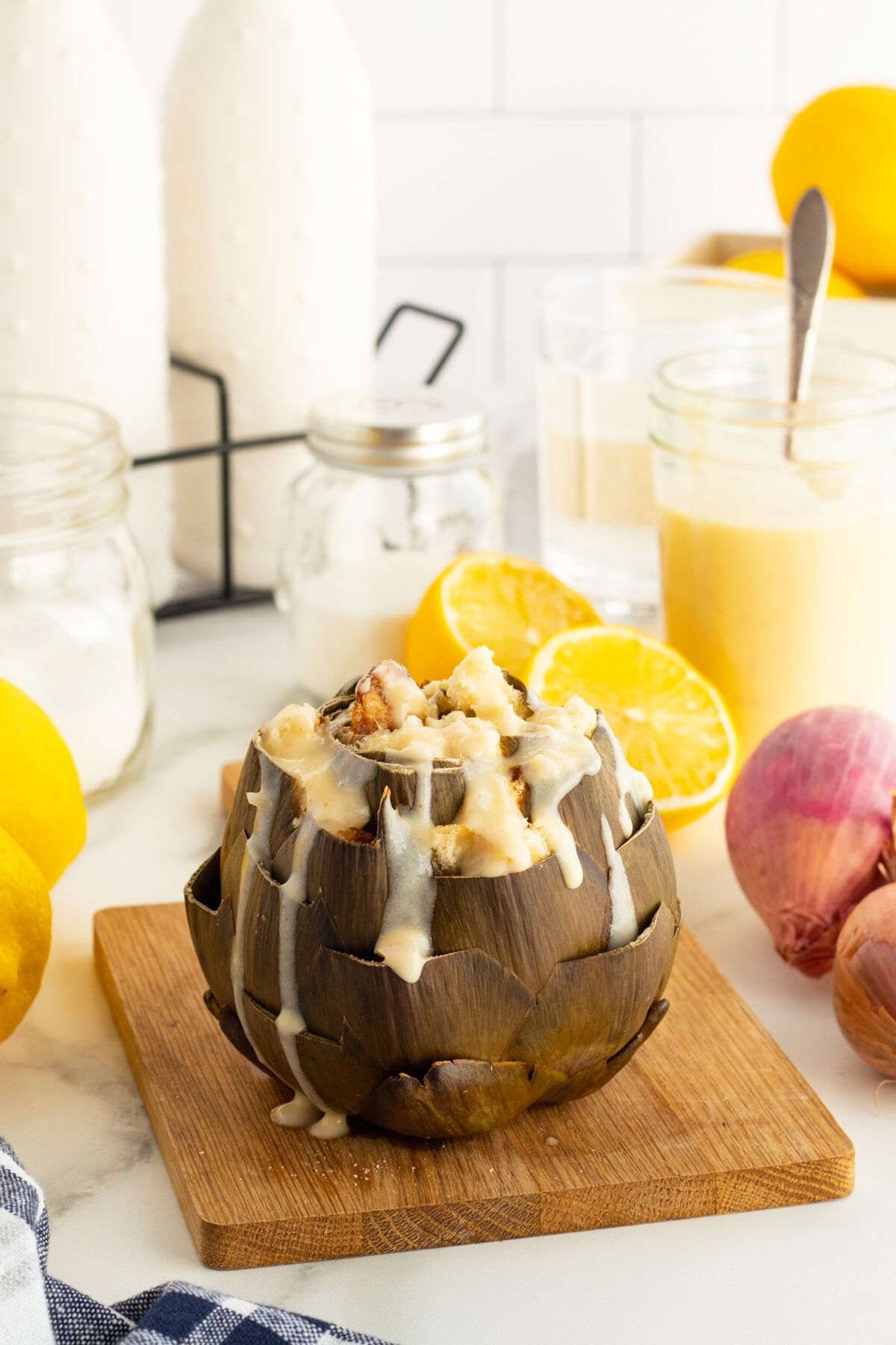 A cooked artichoke stuffed with a creamy filling sits on a wooden board, drizzled with sauce. Surrounding it are lemons, shallots, glass jars, and a glass of sauce, all on a white kitchen counter.