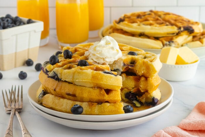 A stack of blueberry waffles topped with whipped cream sits on a plate, with a pat of butter, fresh blueberries, two forks, and glasses of orange juice in the background.