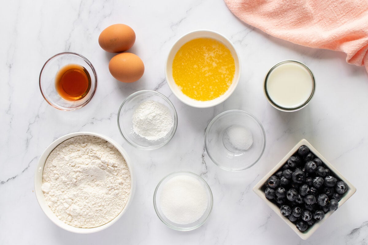 A flat lay of baking ingredients on a marble surface: two eggs, vanilla extract, melted butter, milk, flour, sugar, baking powder, salt, and a bowl of fresh blueberries, with a peach towel in the corner.