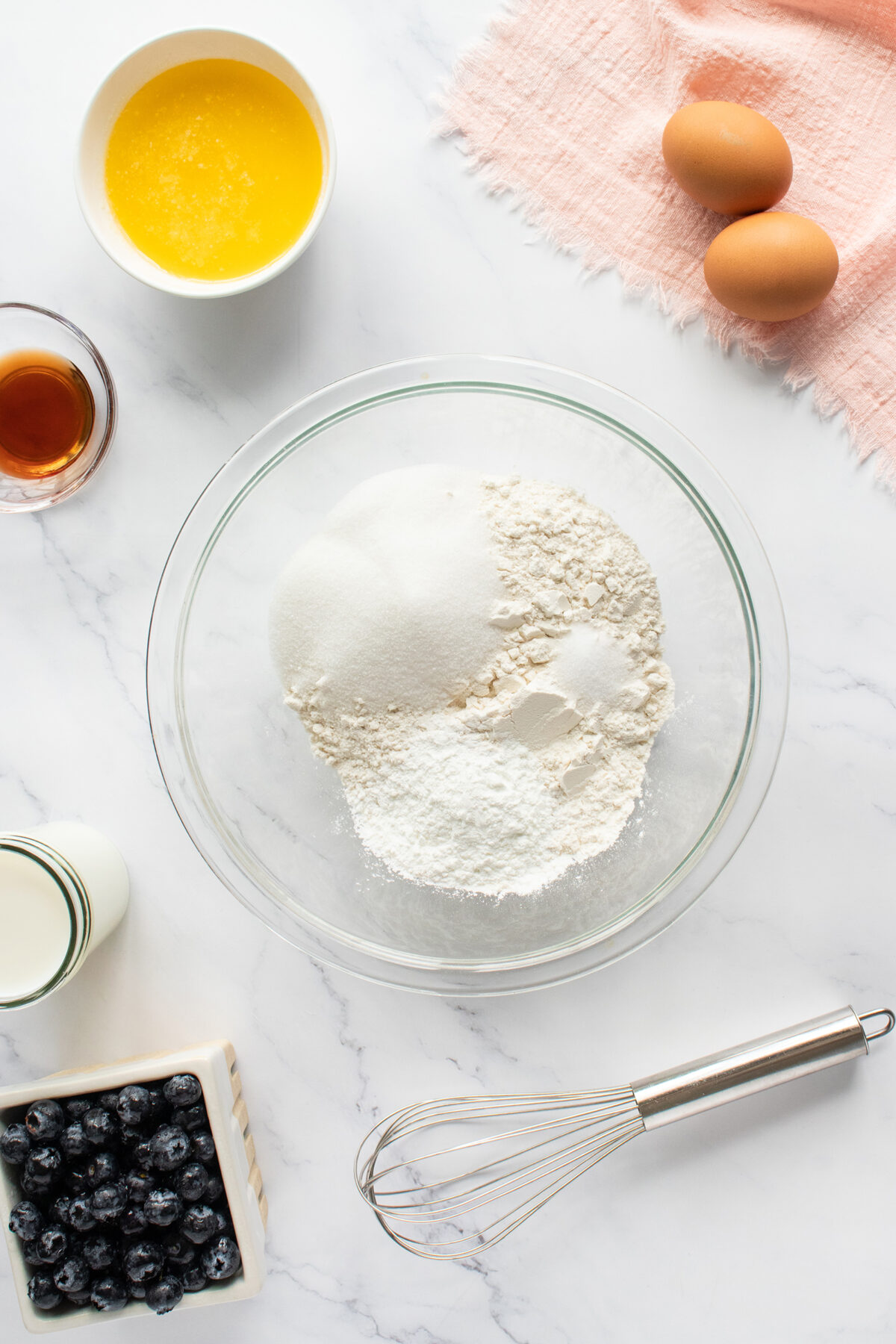 A glass bowl with flour and sugar, surrounded by eggs, melted butter, vanilla, milk, blueberries, and a whisk on a marble surface with a pink cloth.