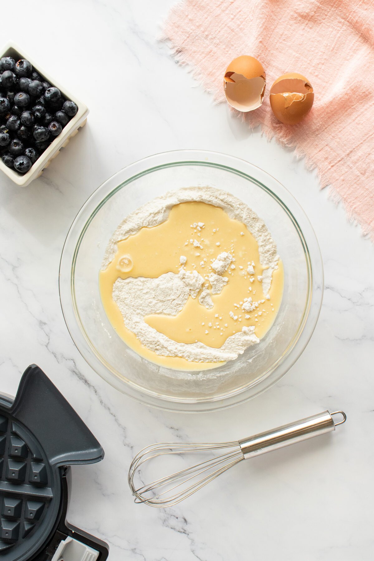 A glass bowl with flour and wet ingredients sits on a marble counter beside a whisk, two cracked eggs, a bowl of blueberries, a pink cloth, and part of a waffle maker.