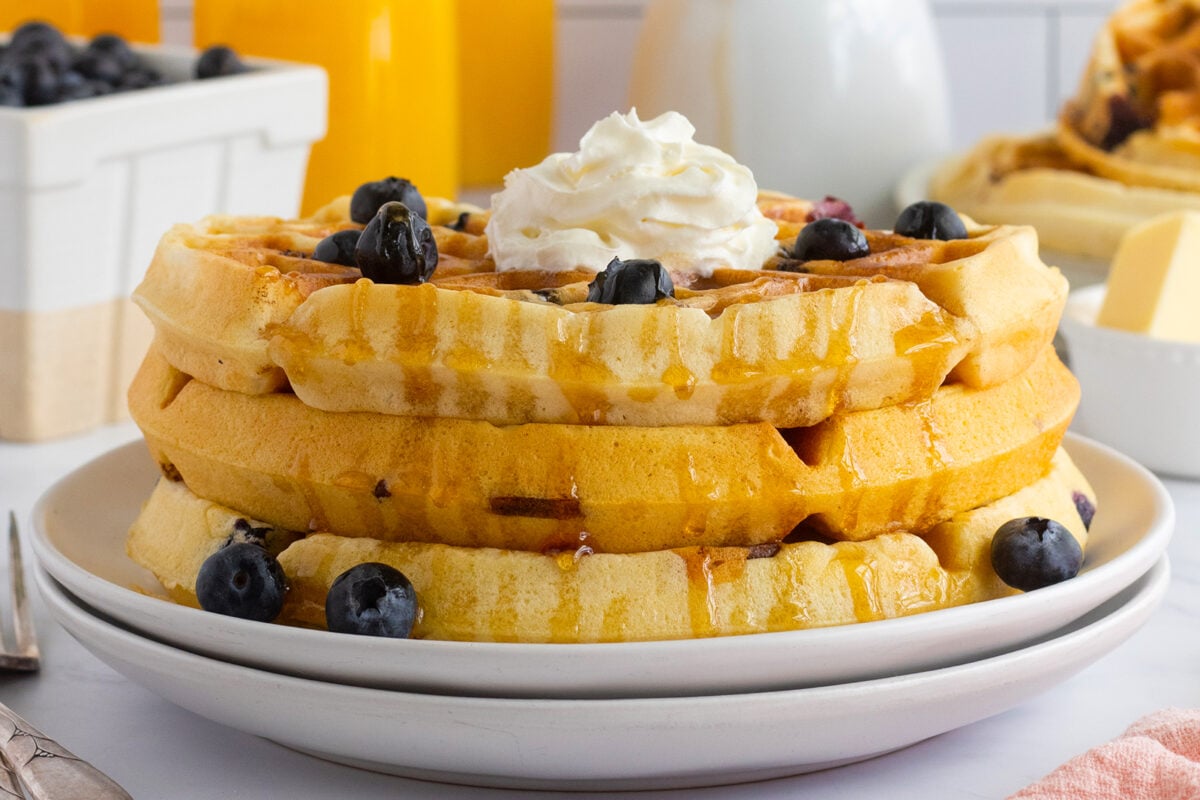 A stack of waffles topped with whipped cream, fresh blueberries, and syrup sits on a white plate, with a fork and butter in the background.
