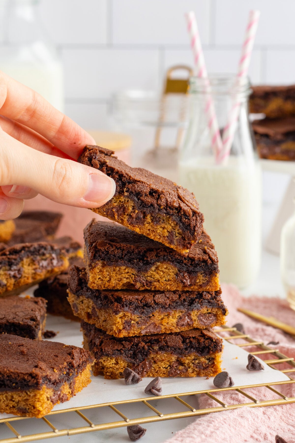 A hand lifts a chocolate chip cookie and brownie bar from a stack of dessert bars on a cooling rack, with milk in glass bottles and straws in the background.