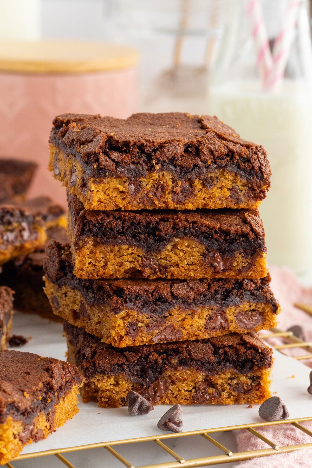 A stack of chocolate chip cookie and brownie bars sits on a cooling rack, showing distinct layers of golden cookie dough and rich chocolate brownie, with chocolate chips visible throughout. Milk bottles are blurred in the background.