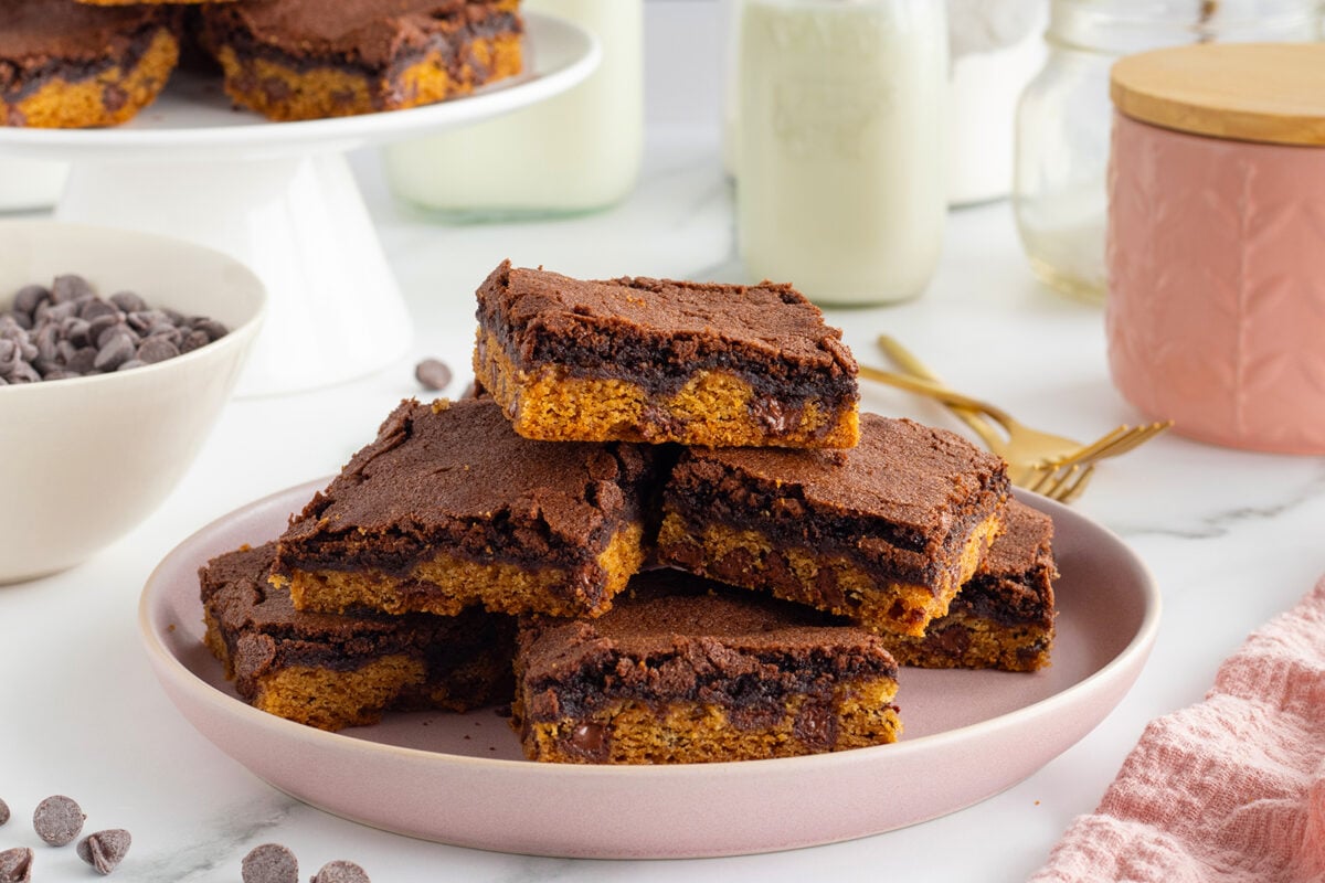 A plate of chocolate-topped cookie bars stacked on a light pink plate, with chocolate chips scattered nearby and milk bottles in the background. Gold forks and additional bars are also visible.