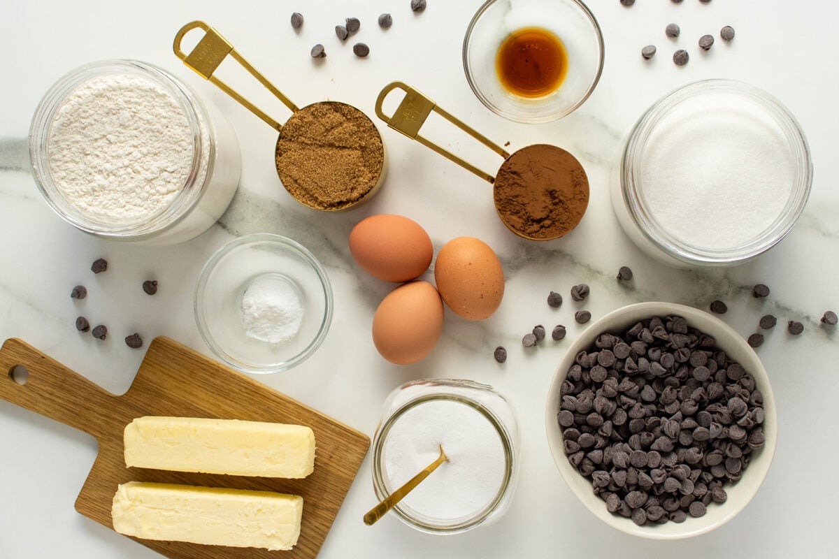 A top view of baking ingredients, including flour, cocoa powder, brown and white sugar, chocolate chips, eggs, vanilla extract, butter, and baking soda, arranged on a white surface.